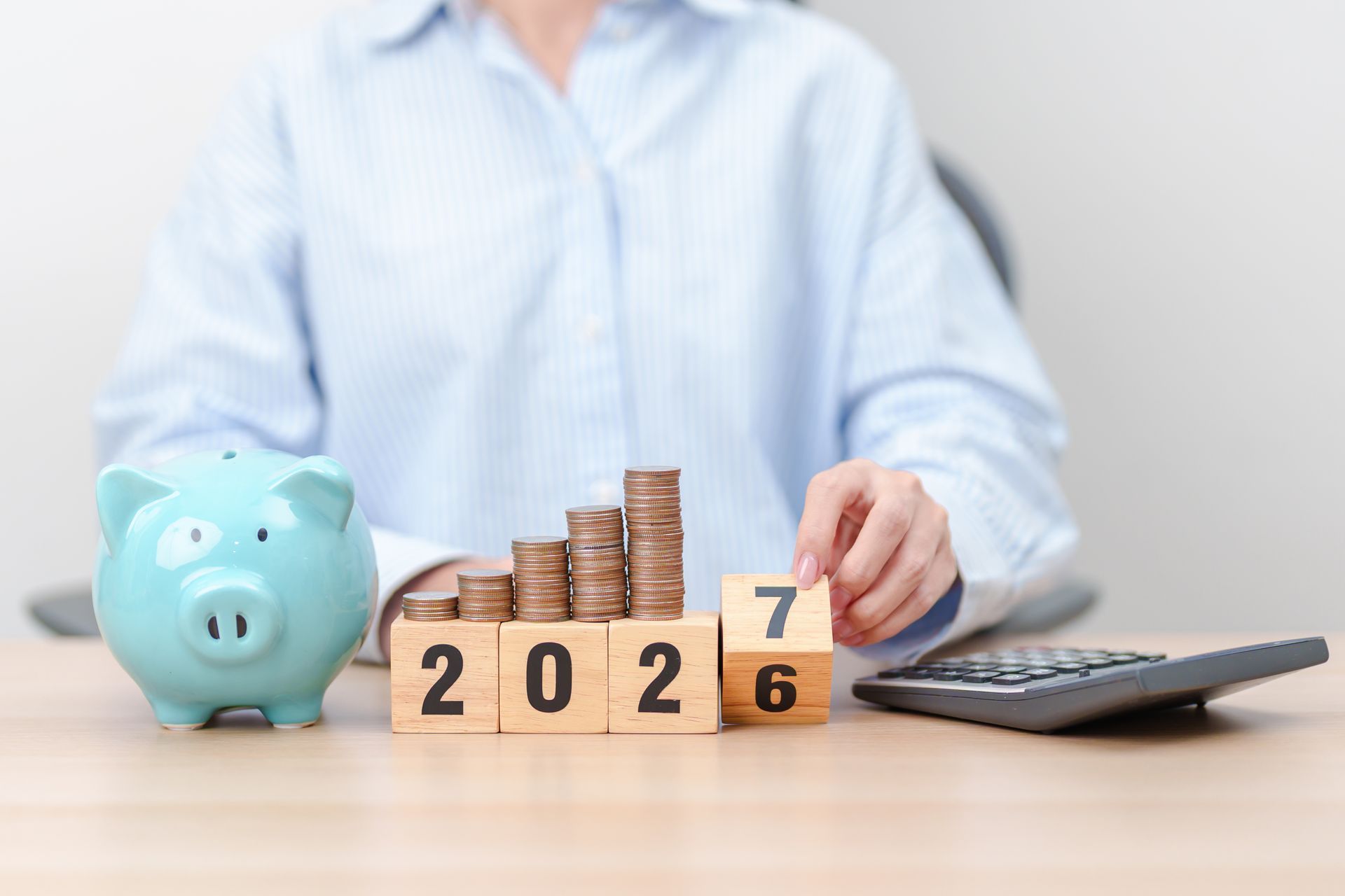 Person at desk with piggy bank, coins, 2026 blocks, and calculator, suggesting savings and budgeting