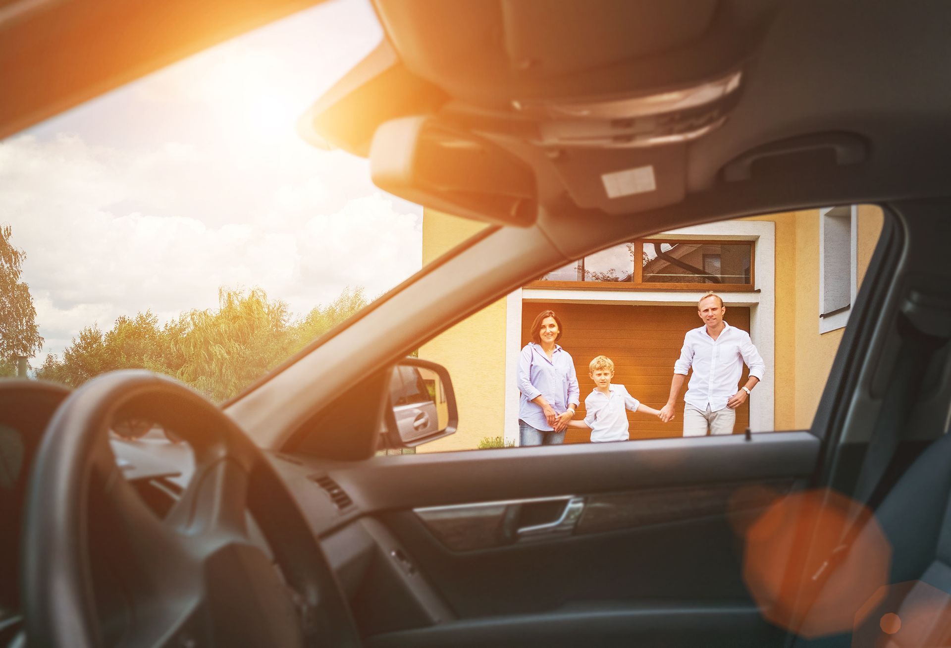 Family standing at a house doorway, seen from inside a car at sunset.