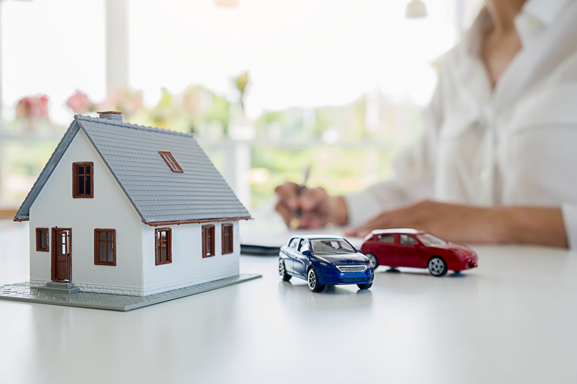 Miniature house model with two toy cars on a desk, with a blurred person in the background