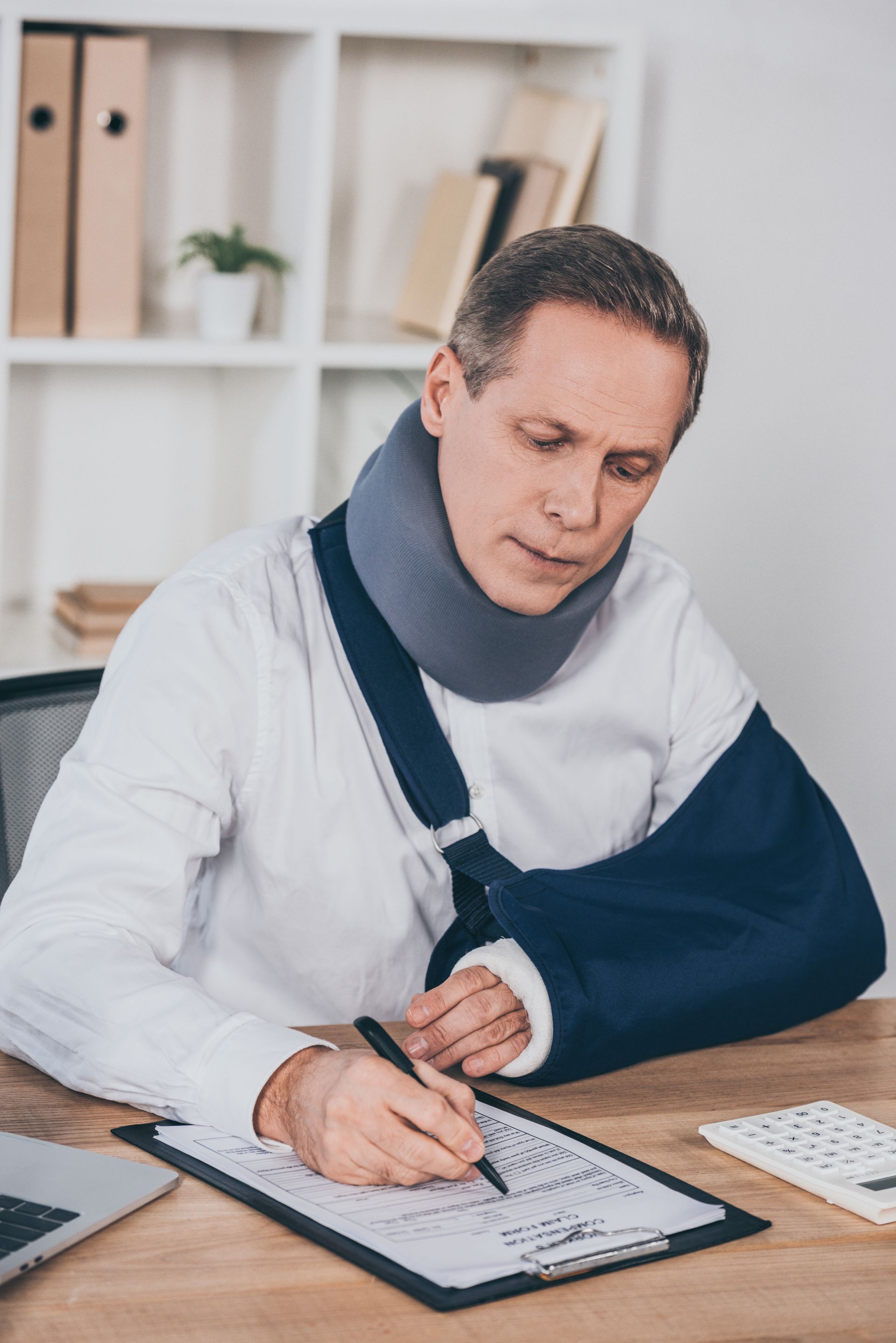 Man with a neck brace writing on a clipboard at a desk in a bright office