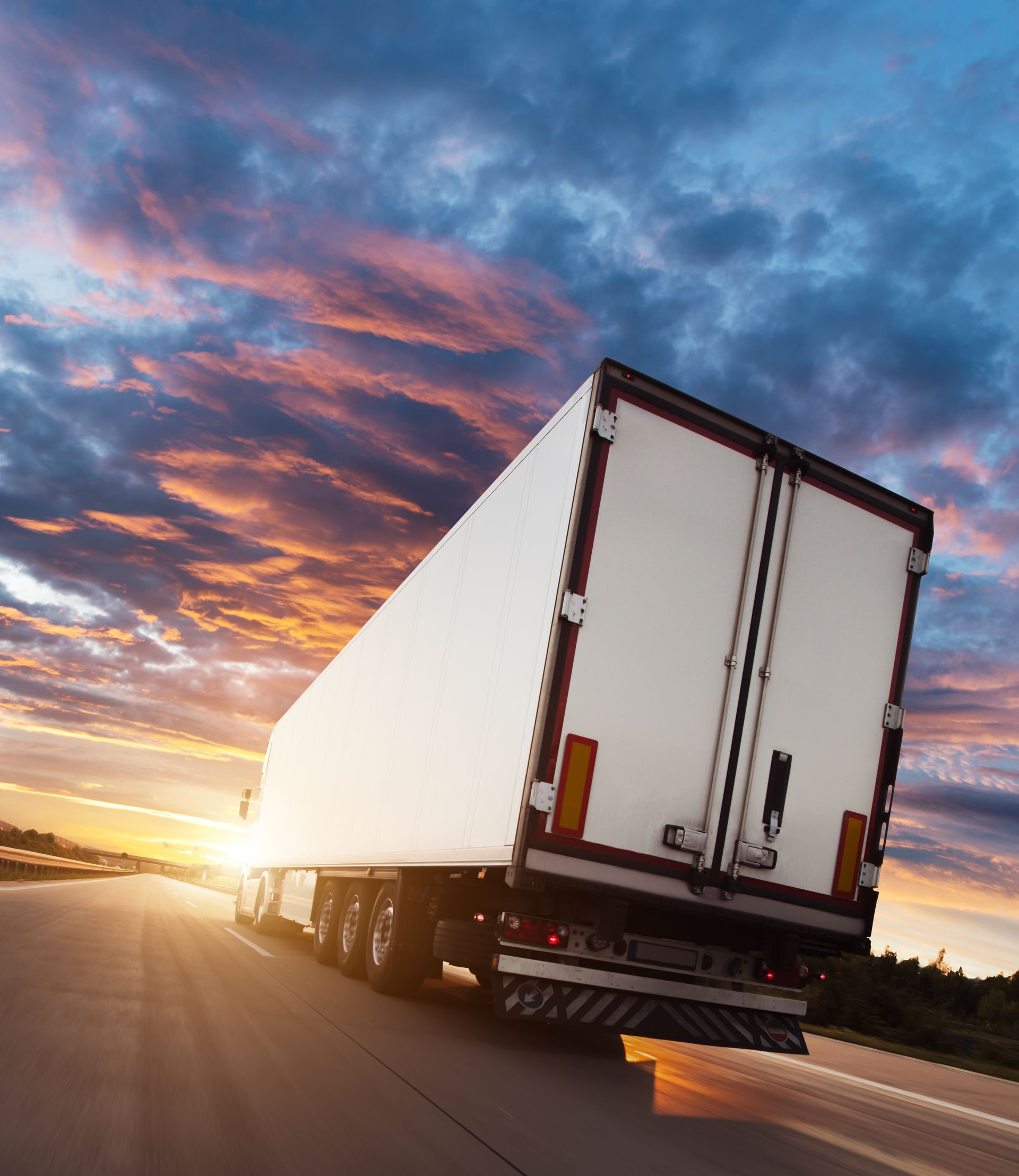 White semi-truck trailer driving on a highway at sunset under a colorful sky