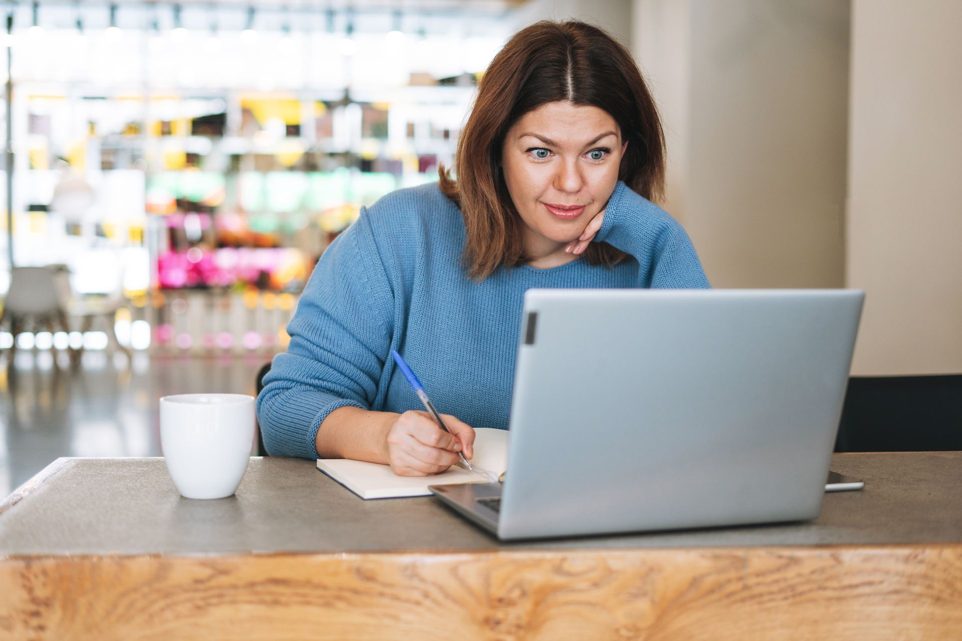 Woman writing beside a laptop at a table in a bright café, with a coffee mug nearby.