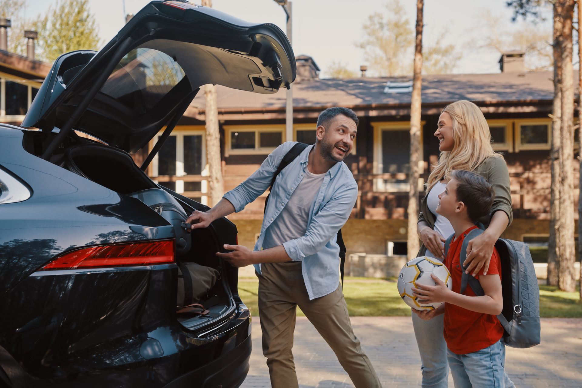 Family unloading bags from a black SUV in a driveway, smiling outside a house.