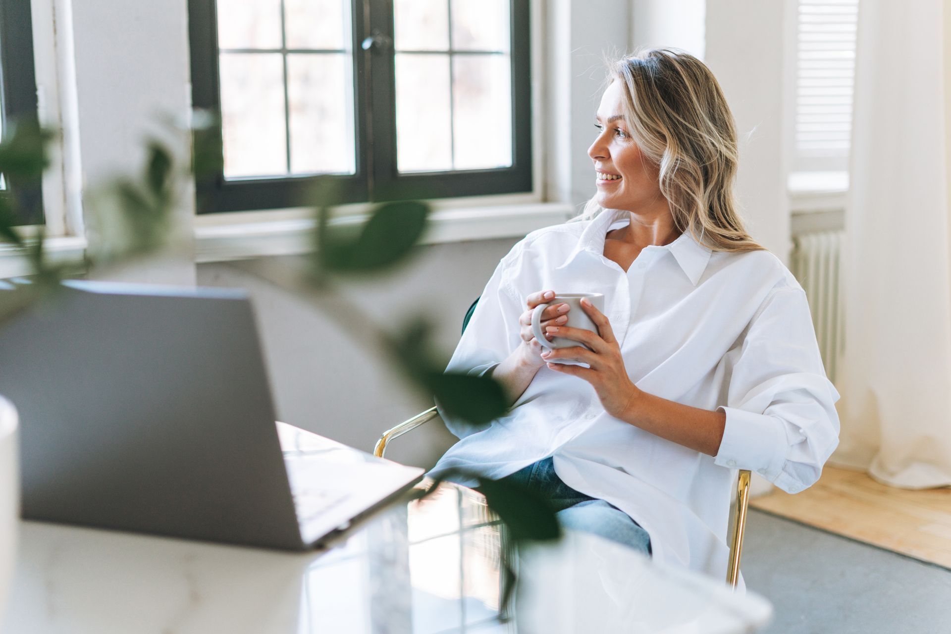 Woman sitting with a laptop and coffee in a bright office, looking out the window.