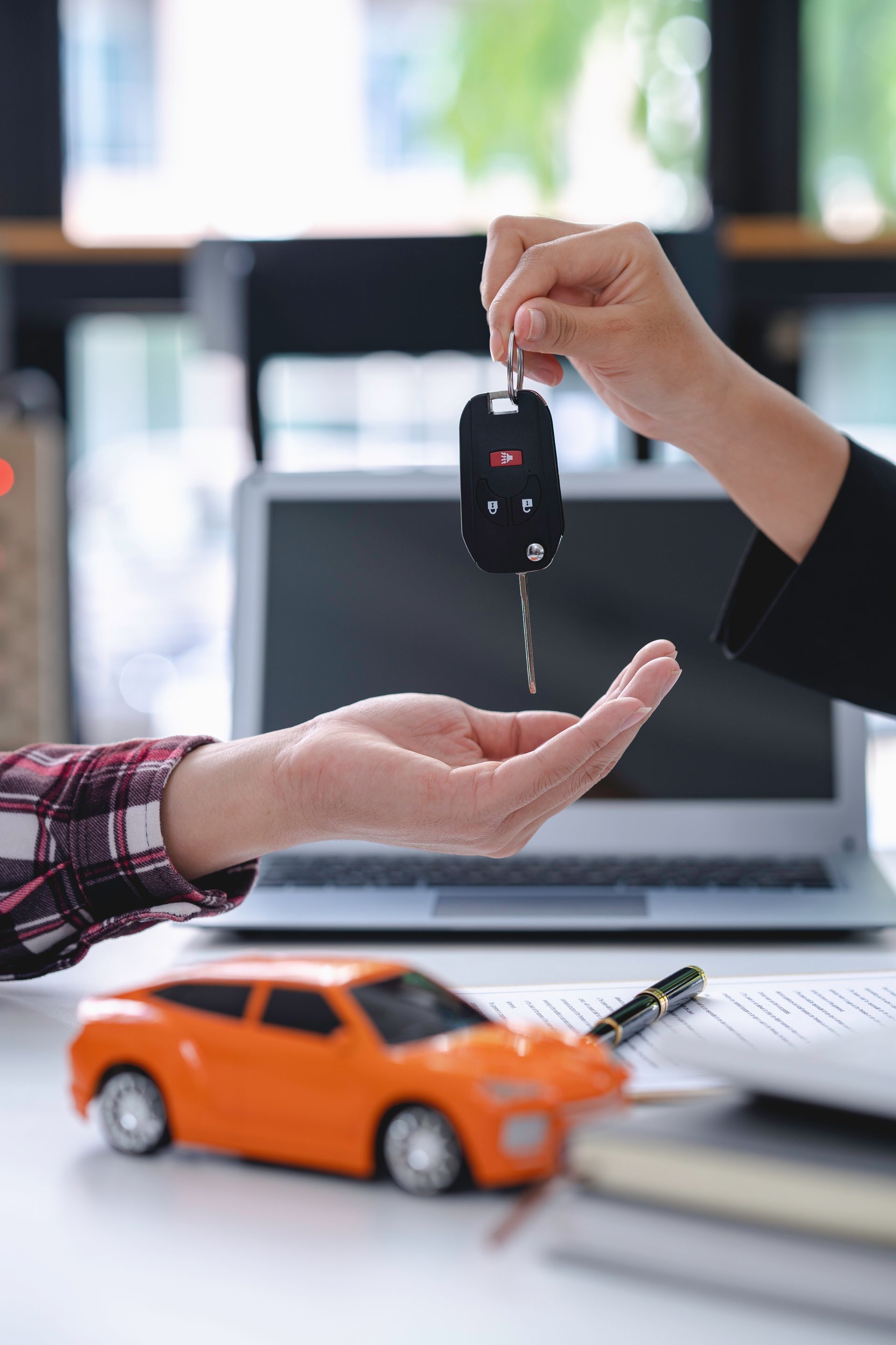 Handing over car keys above an orange toy car beside a laptop on a desk