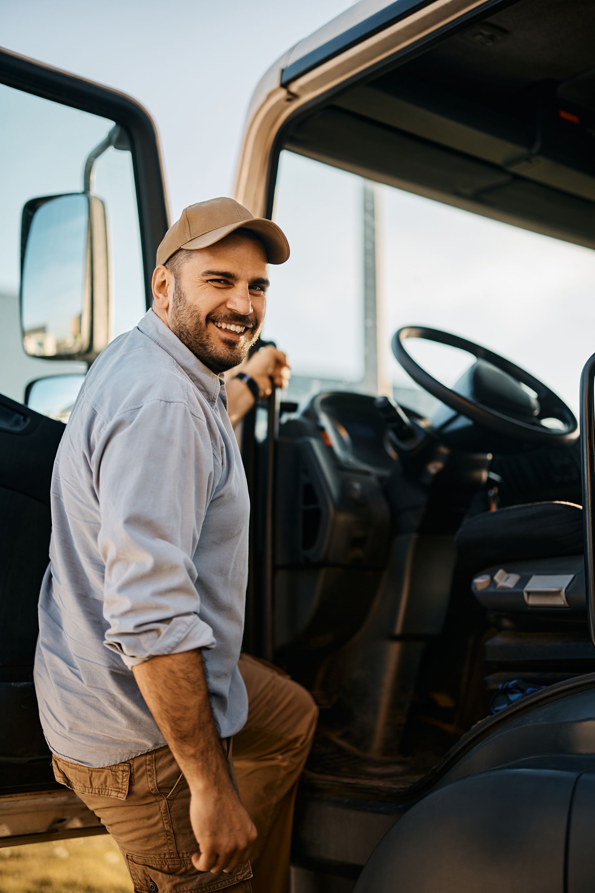 Smiling person standing beside an open truck door, wearing a cap and light shirt.