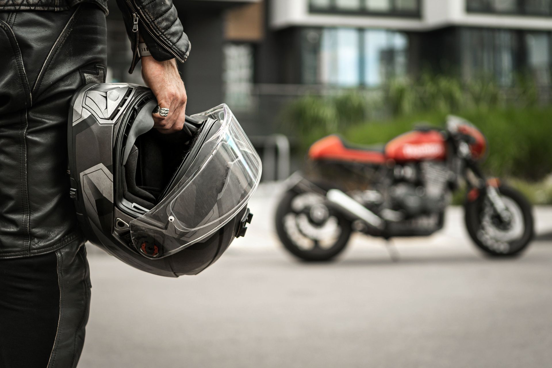 Person in black motorcycle gear holding a helmet, with a red motorcycle blurred in the background.