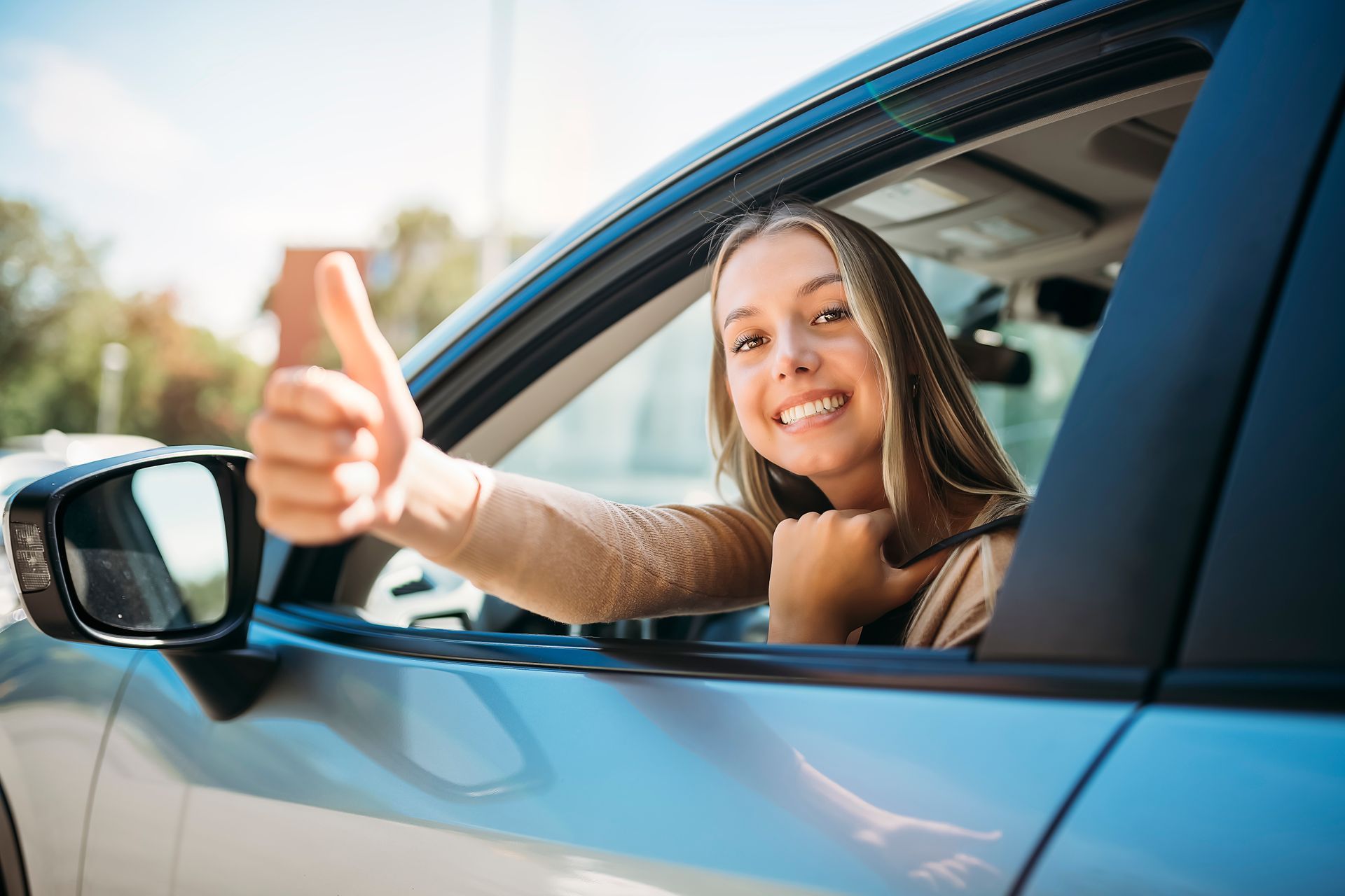 Smiling driver giving a thumbs-up from a blue car window on a sunny day