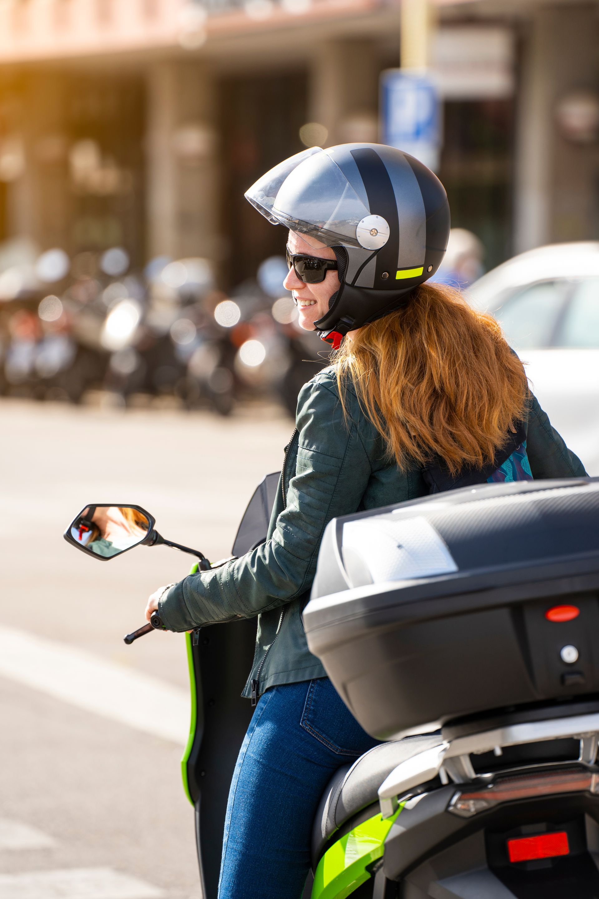 Motorcyclist in a helmet and sunglasses rides through a sunlit street with a top case on the bike