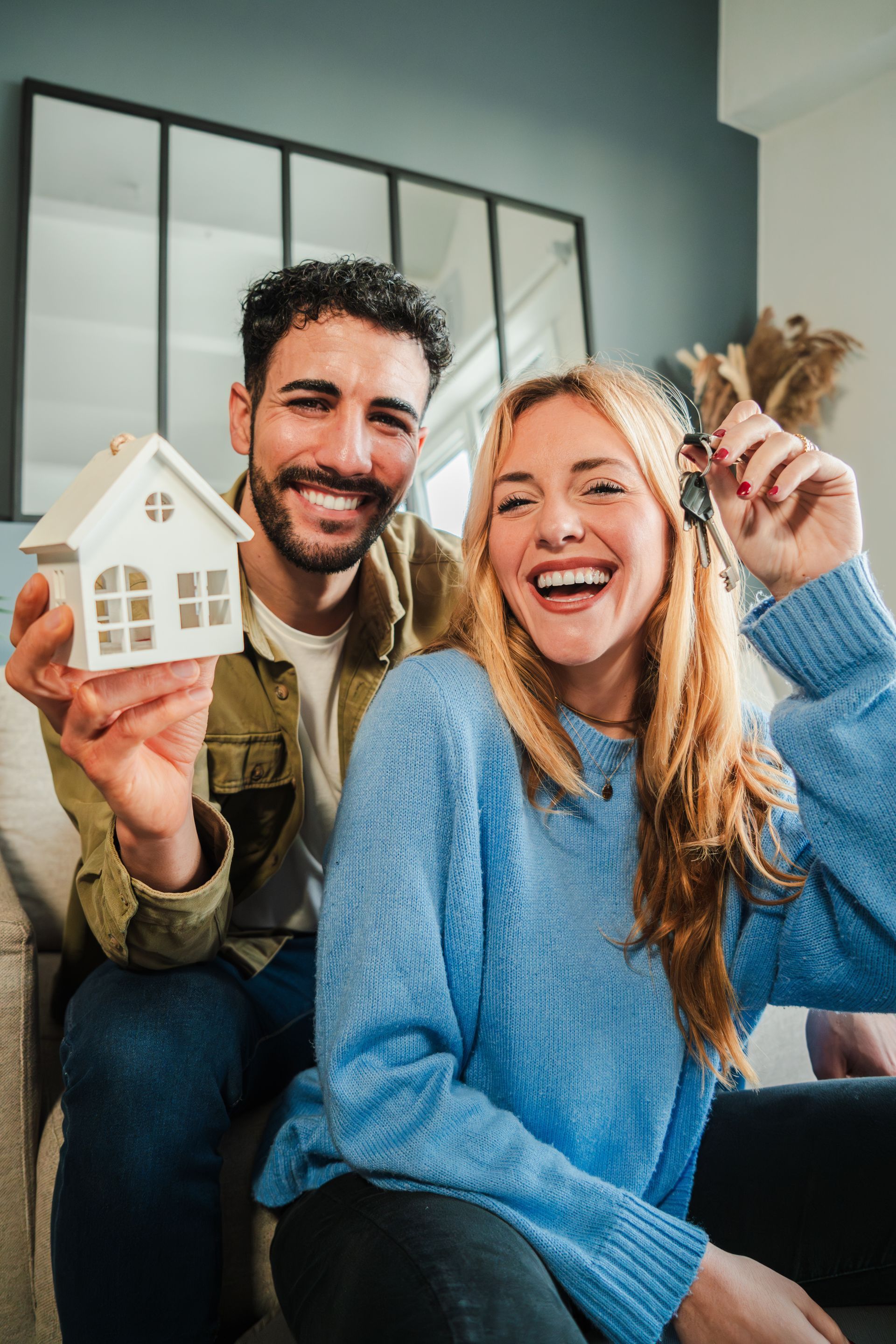 Smiling couple holding a small house model, seated indoors in a living room.