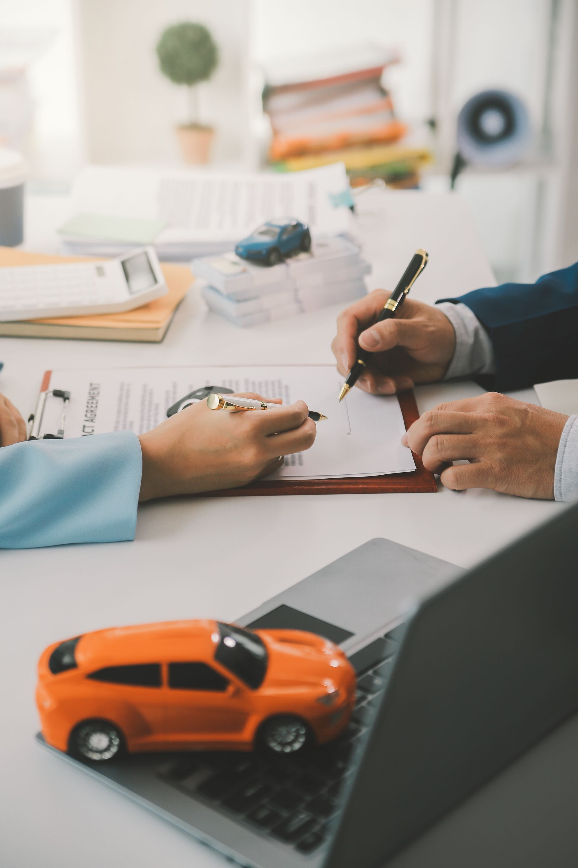 Hands signing a car sale contract beside an orange toy car on a laptop keyboard.