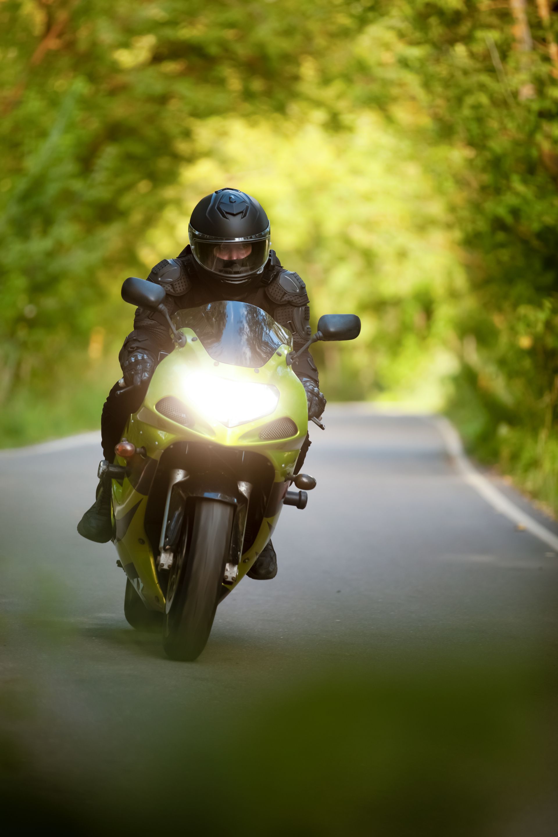 Motorcyclist in black gear riding toward the camera on a winding road, headlights bright against a green forest background