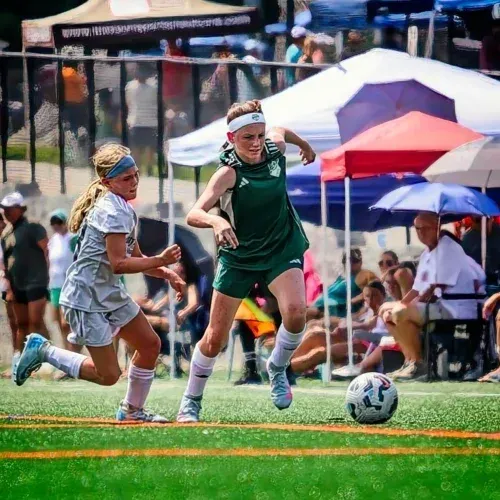 Two soccer players chase the ball on a field, with tents and spectators in the background.
