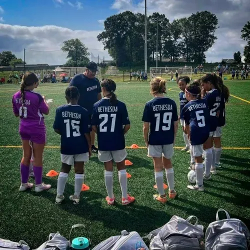 Youth soccer team in navy uniforms standing on the sideline during a match, with cones and gear nearby.