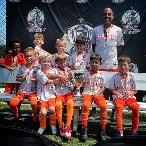 Youth soccer team posing with a trophy on a stage, wearing white jerseys and orange socks.