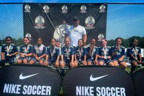 Youth soccer team posing with trophies and coach behind Nike Soccer banners at an outdoor field.