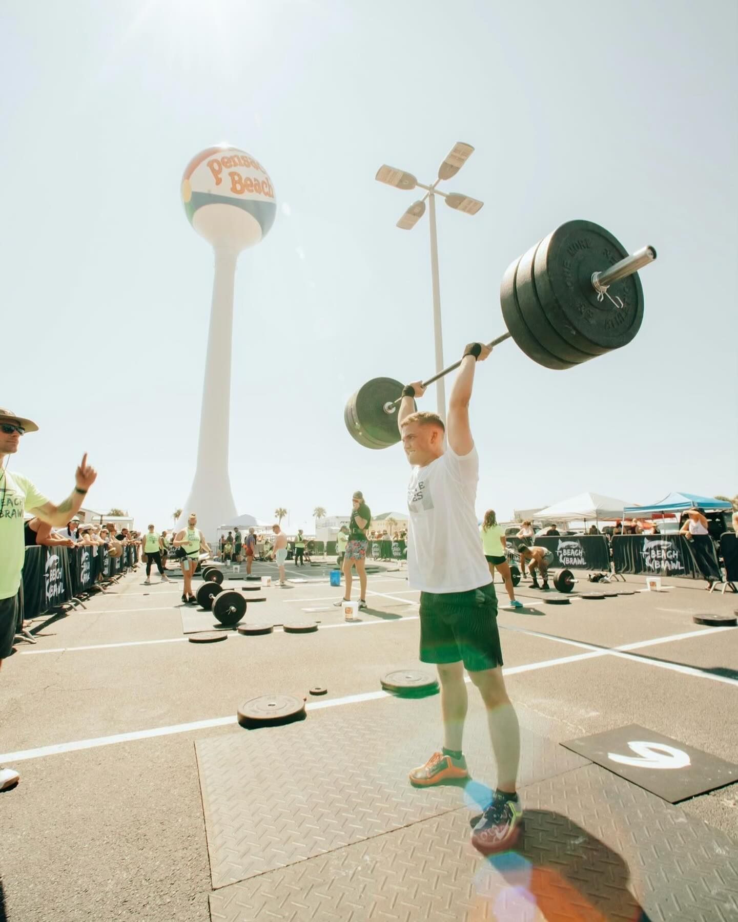 A man is lifting a barbell in a parking lot with a water tower in the background