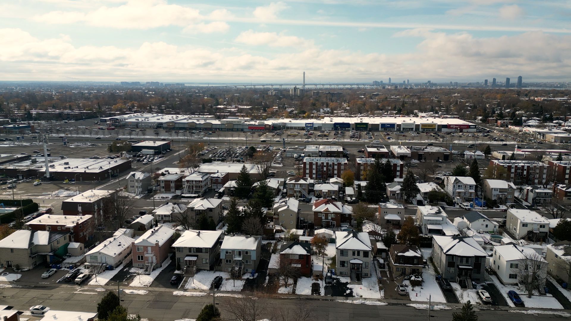 Vue aérienne d'un quartier résidentiel enneigé, avec des bâtiments commerciaux et la silhouette d'une ville au loin.