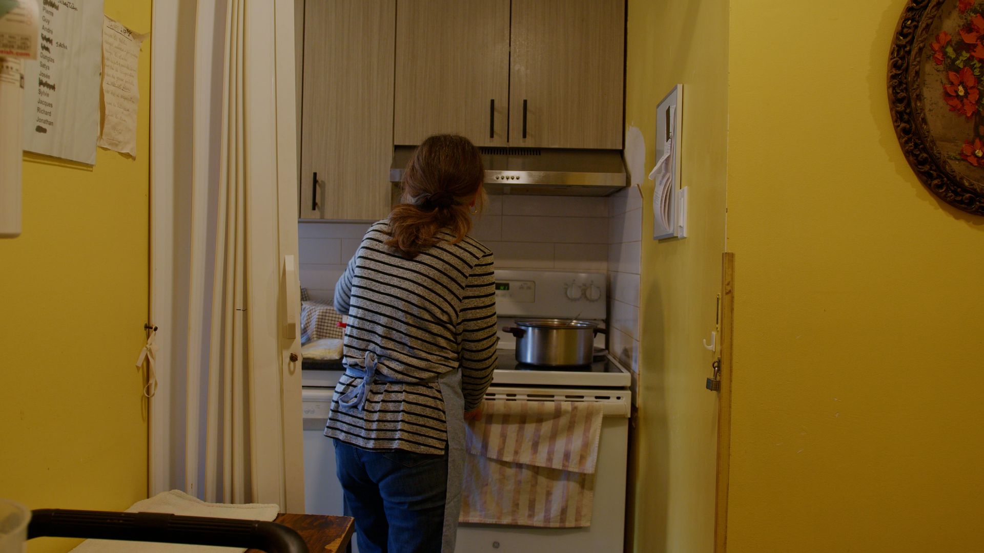 Une femme est assise devant un fourneau dans une cuisine aux murs jaunes. Elle regarde une casserole.