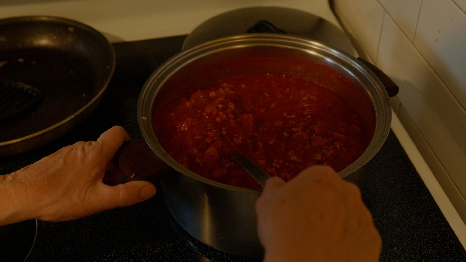 Une personne remuant une casserole de sauce tomate sur une plaque de cuisson, avec une poêle en arrière-plan.