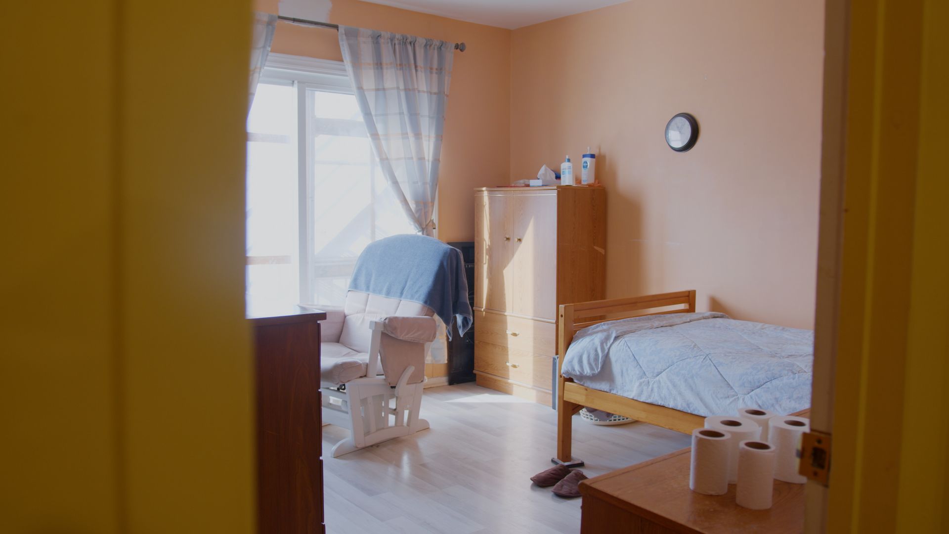 Chambre avec lit, fauteuil à bascule et commode, vue depuis la porte. Murs orange clair, couette bleue.