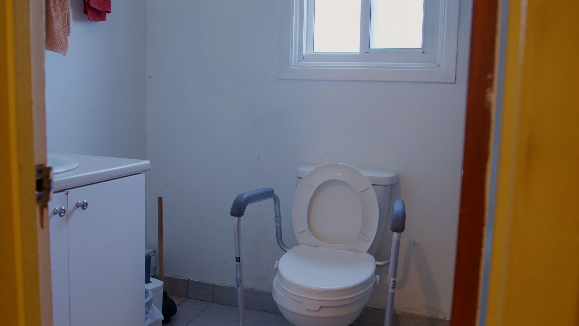 Salle de bains avec siège de toilette surélevé et barres d'appui ; meuble blanc et petite fenêtre.