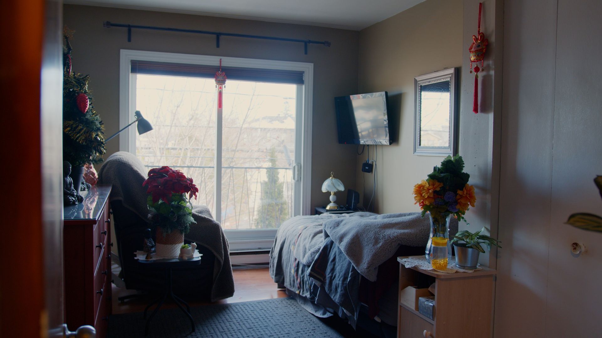 Chambre confortable avec lit, petite télévision, fenêtre, fleurs et décorations. La lumière du soleil y pénètre abondamment.