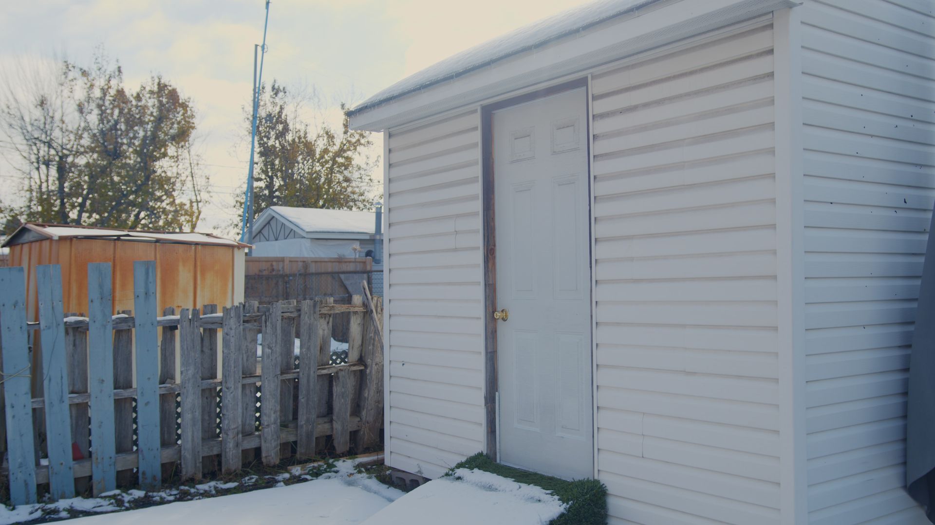 Cabanon blanc à la porte ouverte dans une cour enneigée, à côté d'une clôture en bois usée par les intempéries.
