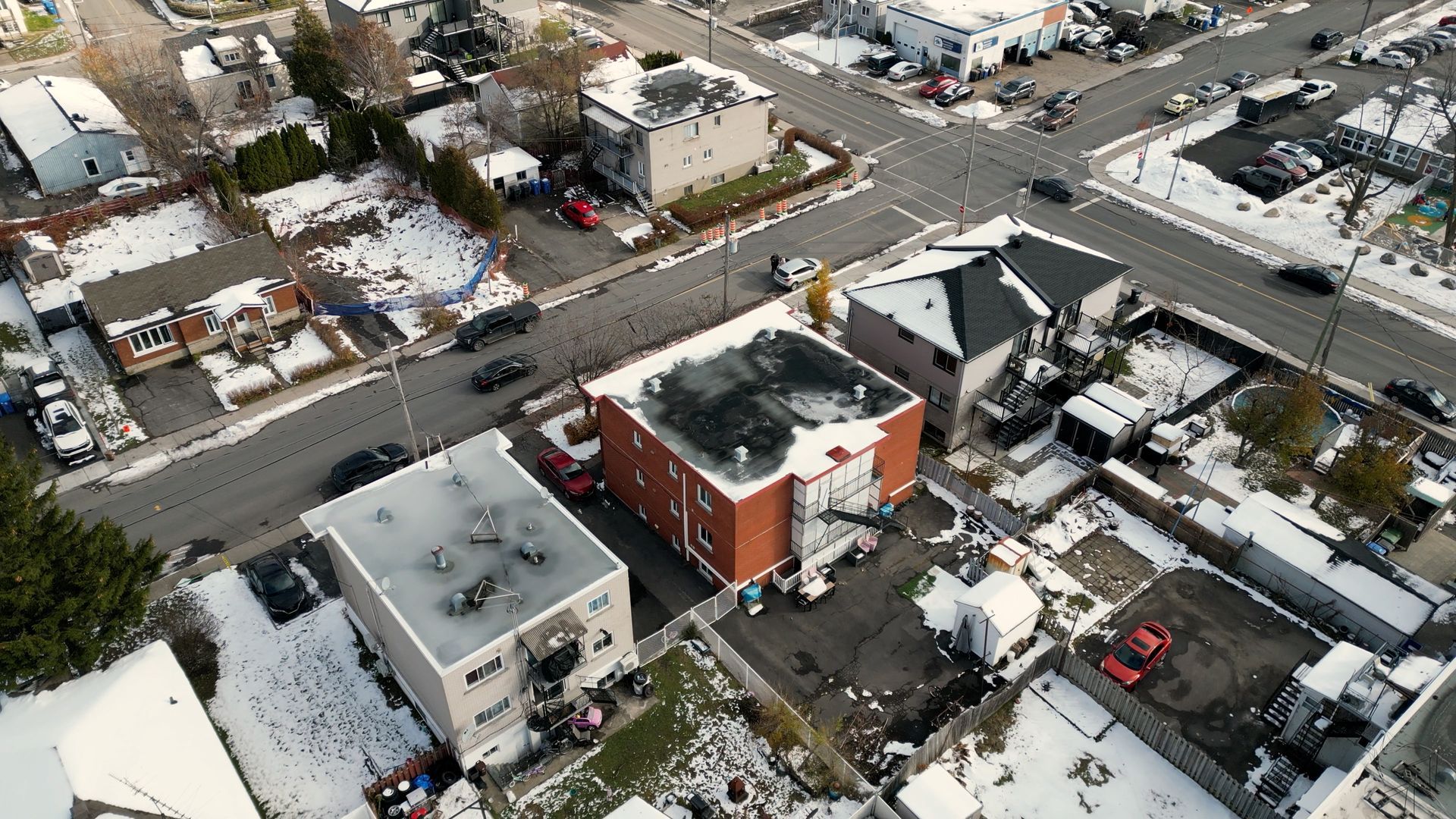 Vue aérienne d'un carrefour enneigé dans un quartier résidentiel, avec des voitures et des bâtiments.