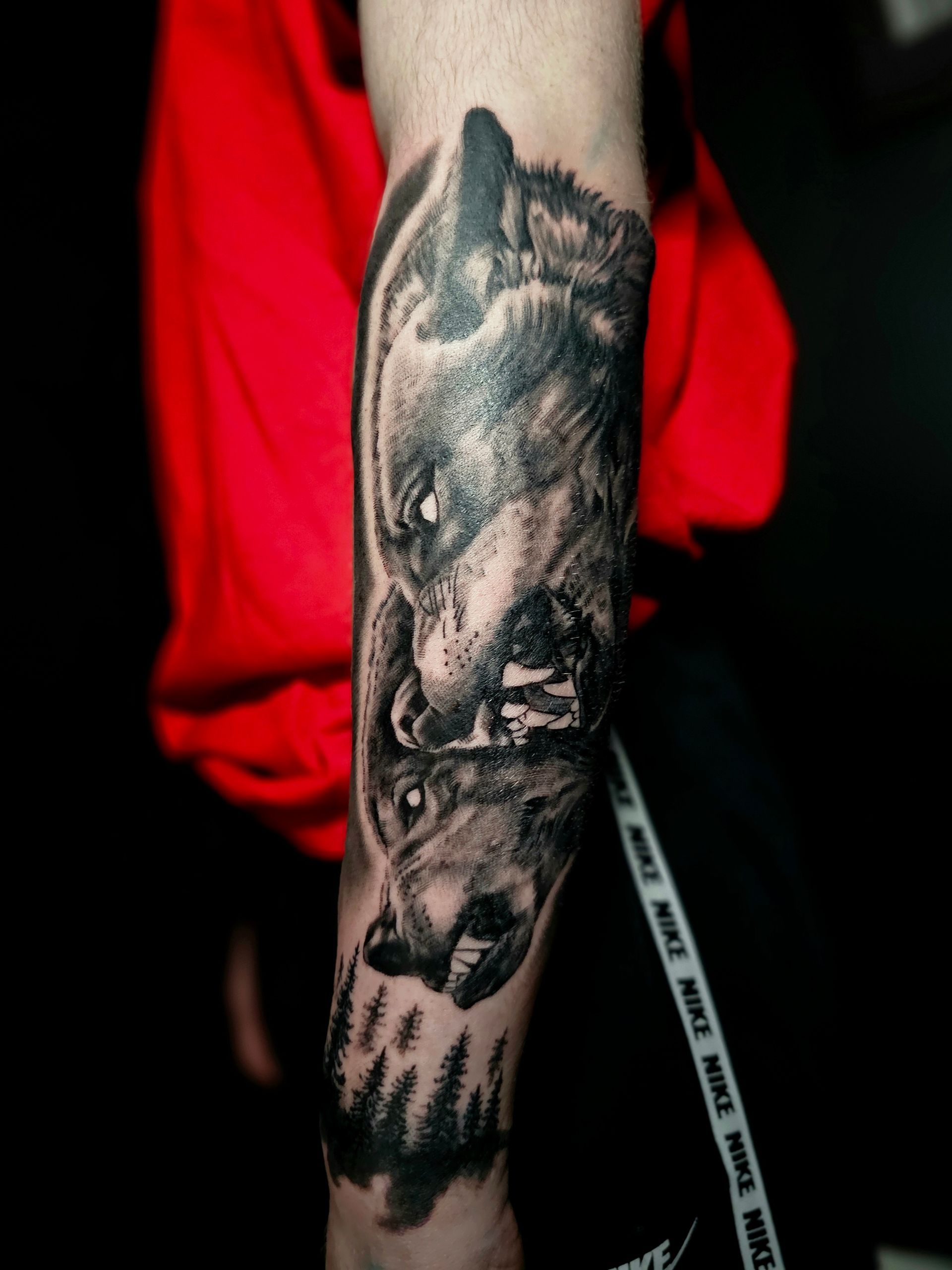 A black-and-grey tattoo on a forearm featuring two snarling wolves above a forest silhouette.