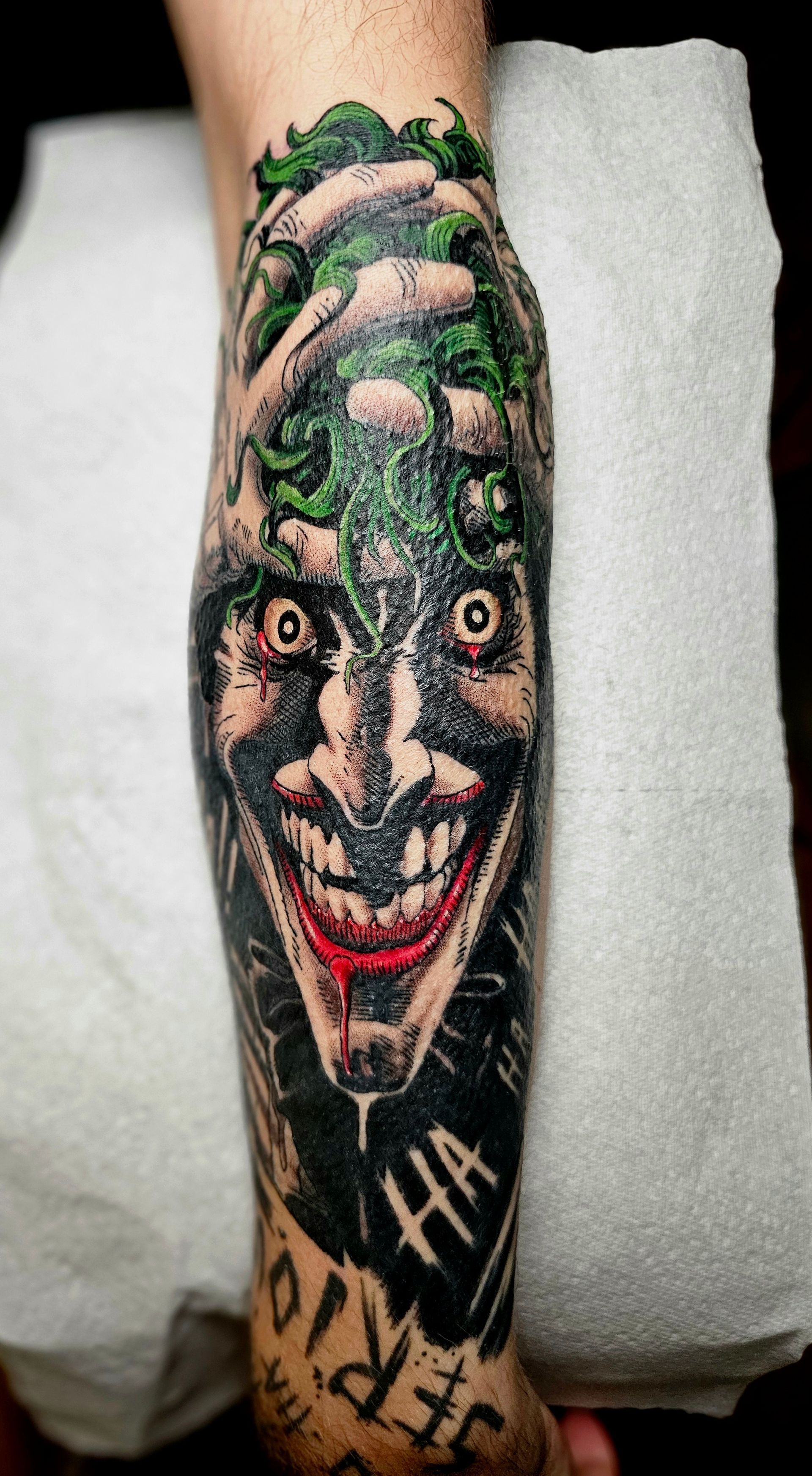 A detailed forearm tattoo of the Joker with wide, staring eyes and a red-lipped grin, surrounded by green hair and text.