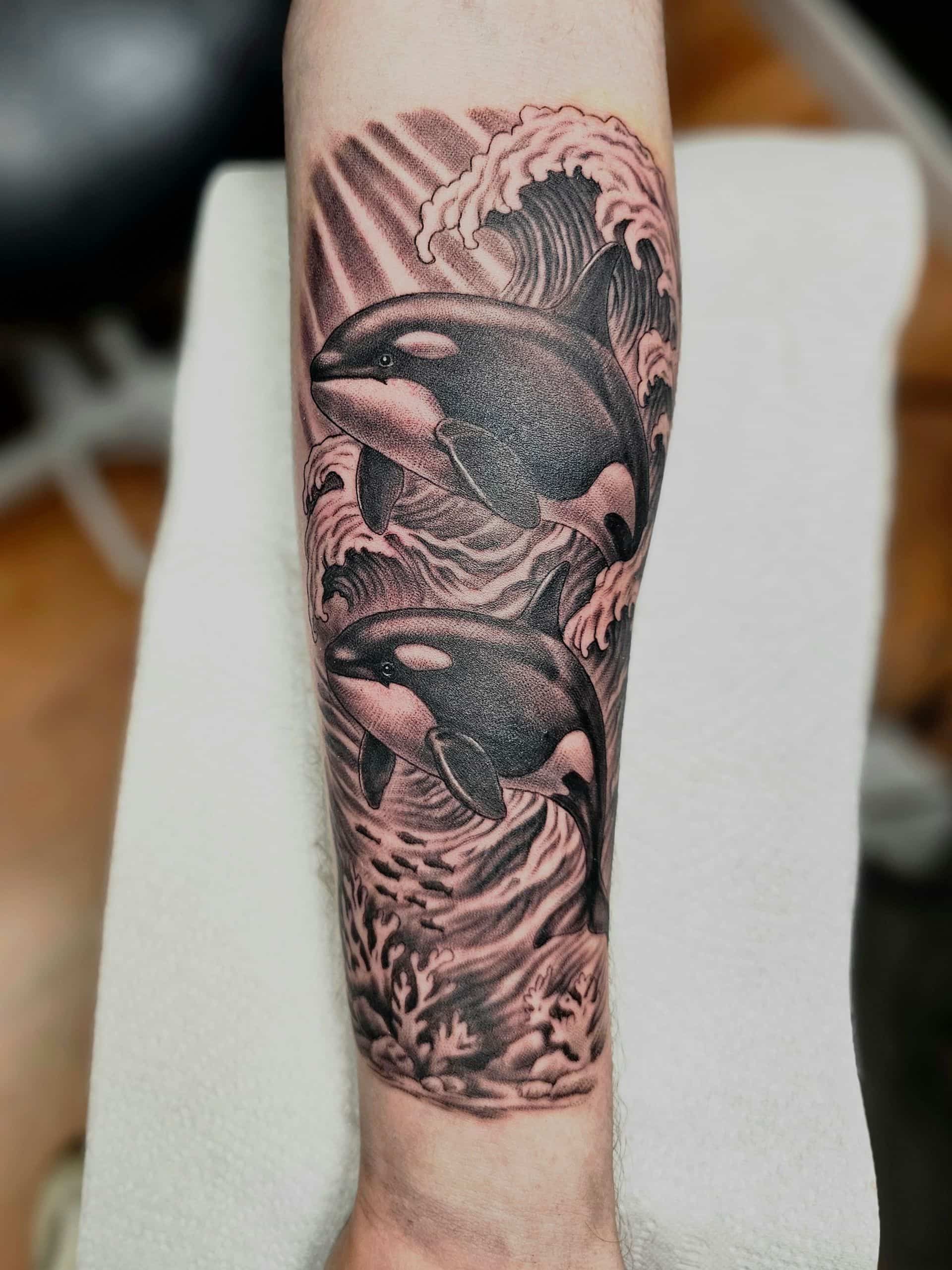 Black and grey forearm tattoo featuring two orcas jumping through crashing ocean waves and light rays.