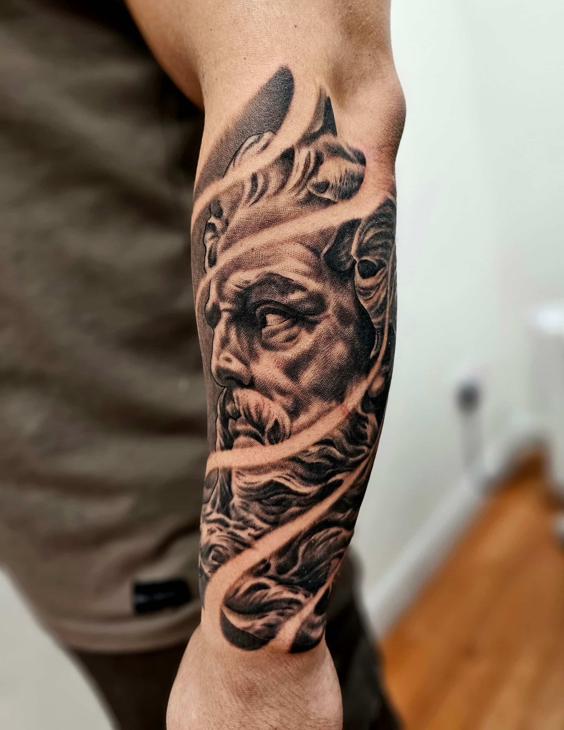 A black-and-grey forearm tattoo of a classical-style bearded face framed by flowing, abstract ribbons.
