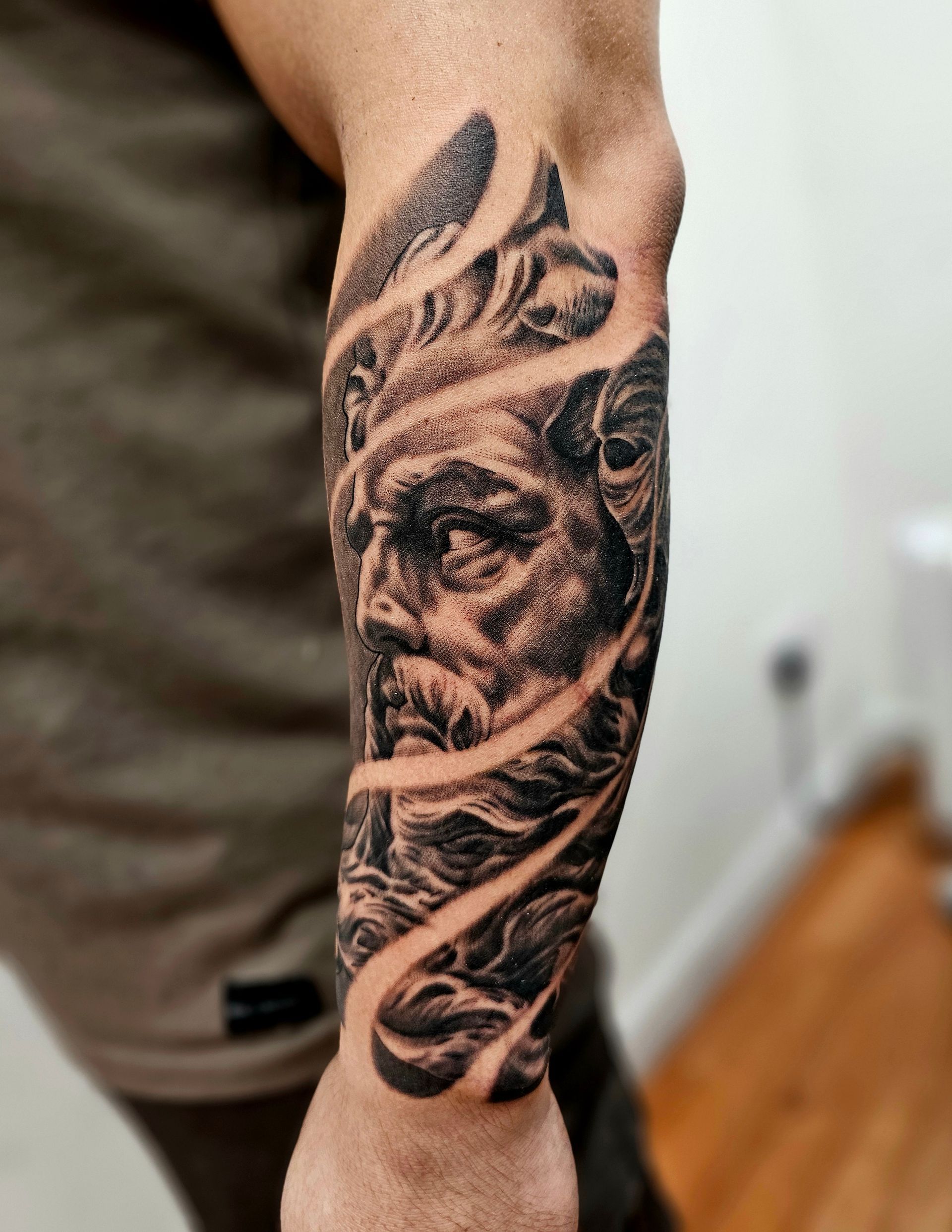 A black-and-grey tattoo on a forearm depicting a classical male bust with flowing hair, stylized by swirling negative space.
