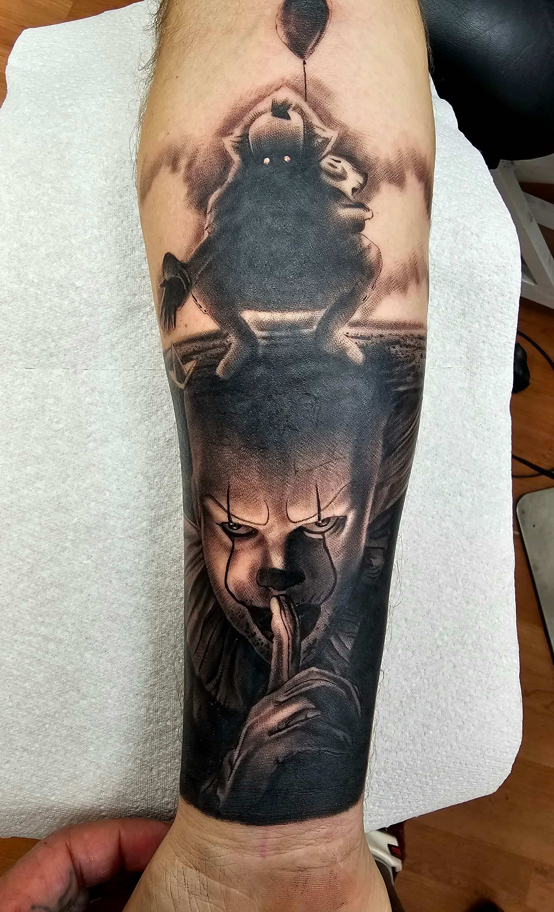 A black and grey tattoo on a forearm depicting Pennywise the clown holding a finger to his lips, with a floating balloon.