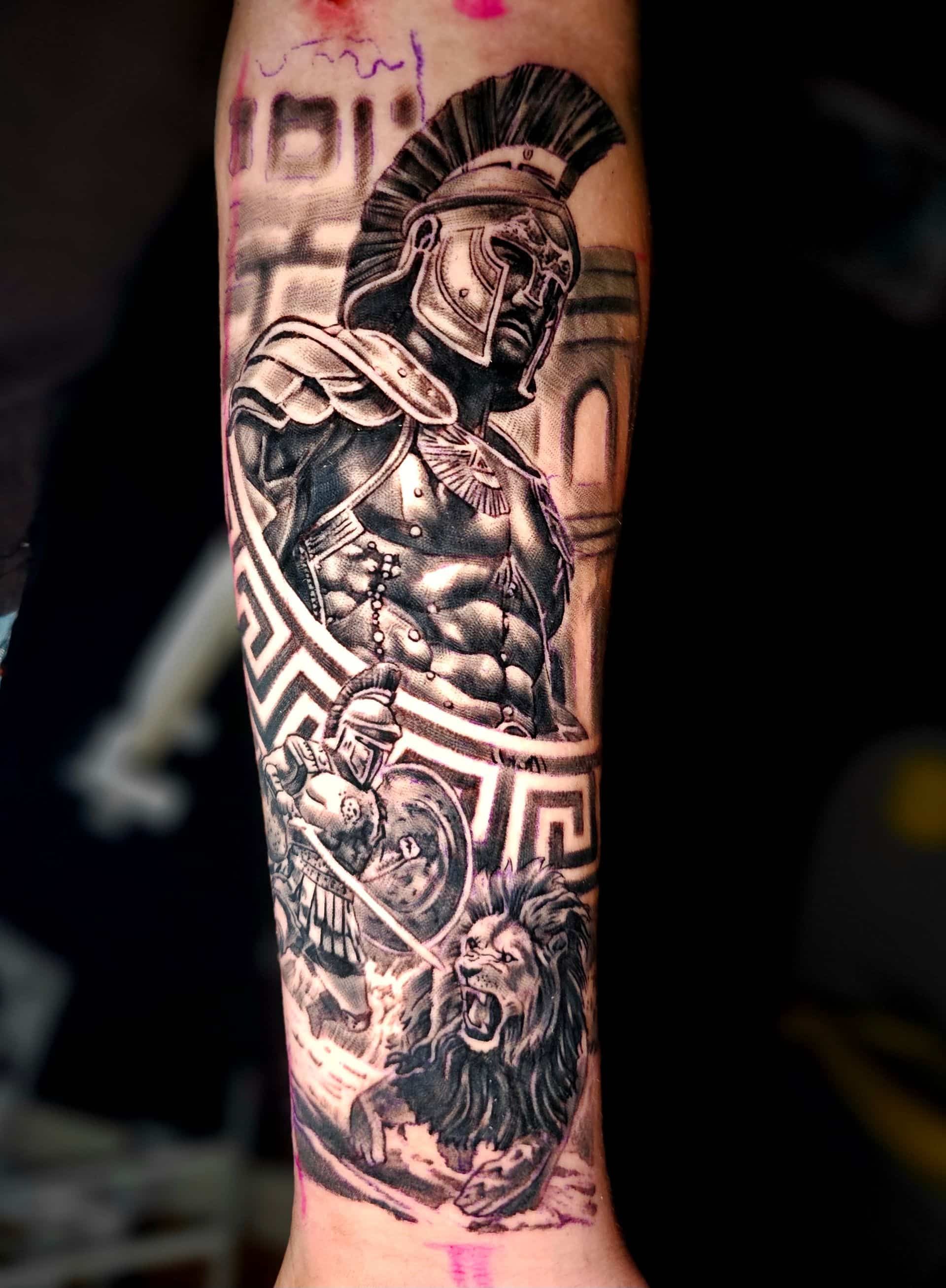 A black and grey tattoo on an arm featuring a Spartan warrior in armor, a Greek key pattern, and a roaring lion.
