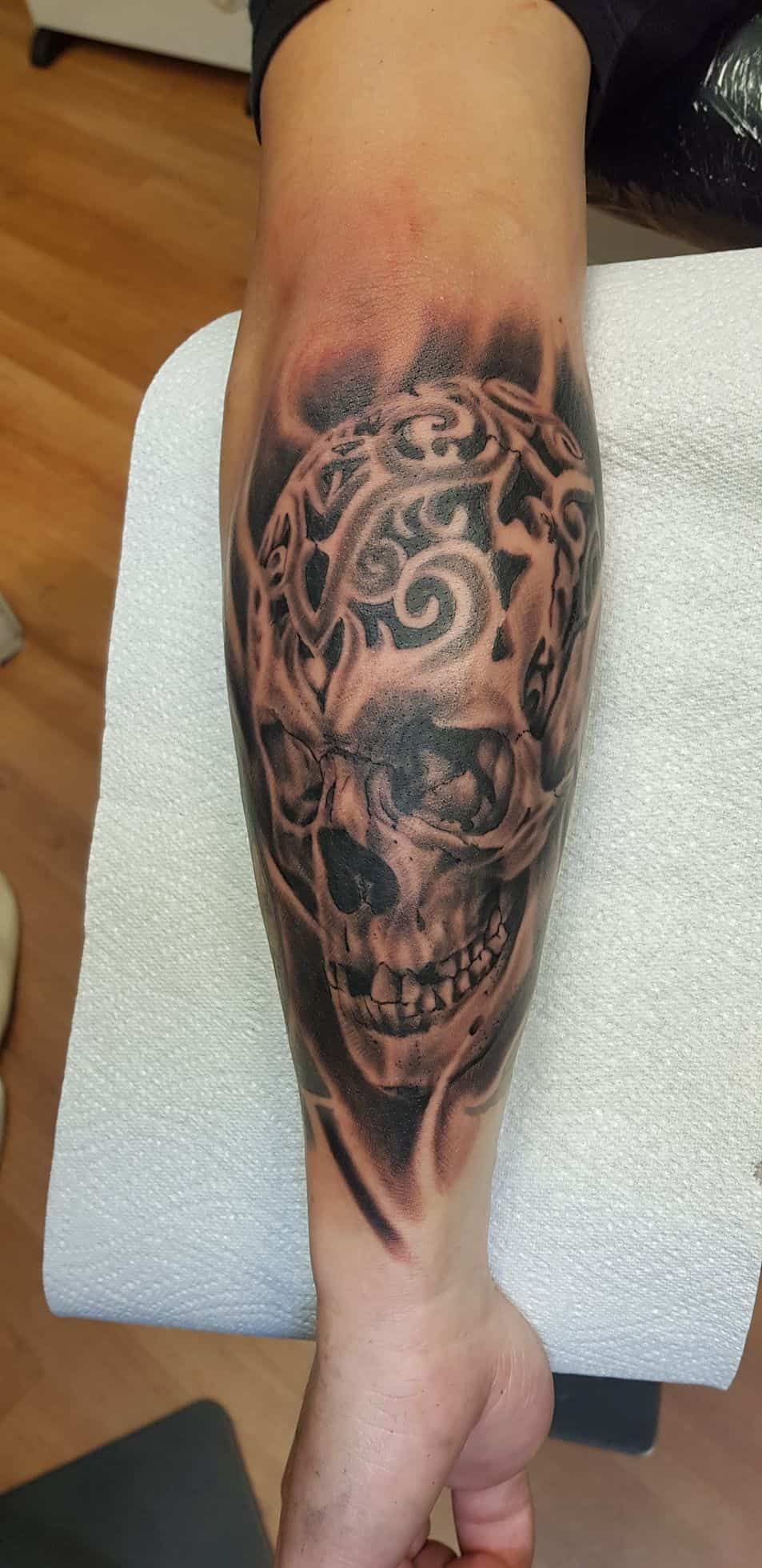 A detailed black and gray tattoo on a forearm featuring a skull adorned with intricate, swirling tribal-style patterns.