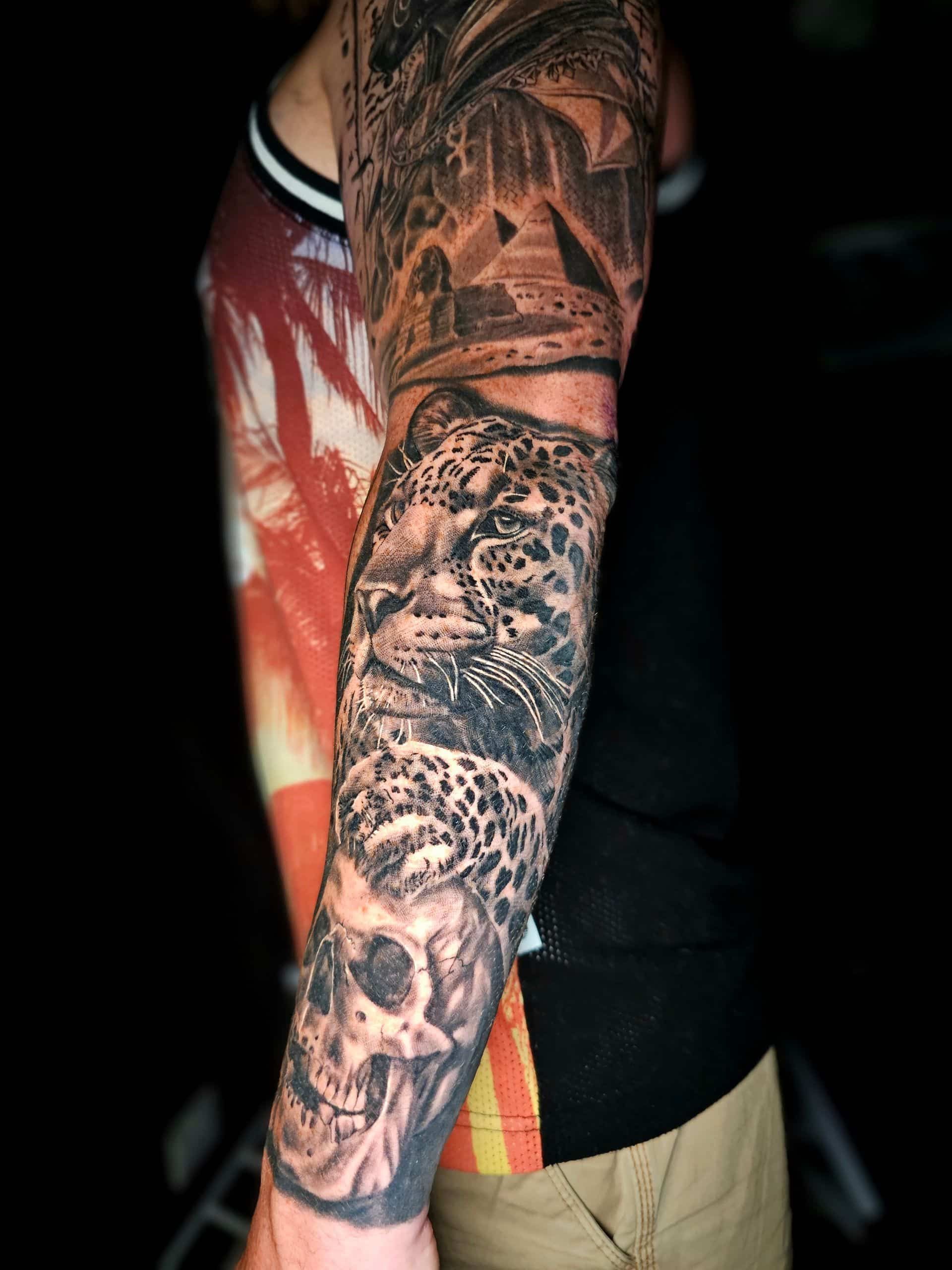 A person’s arm featuring an intricate black and gray tattoo sleeve depicting ancient pyramids, a leopard, and a skull.