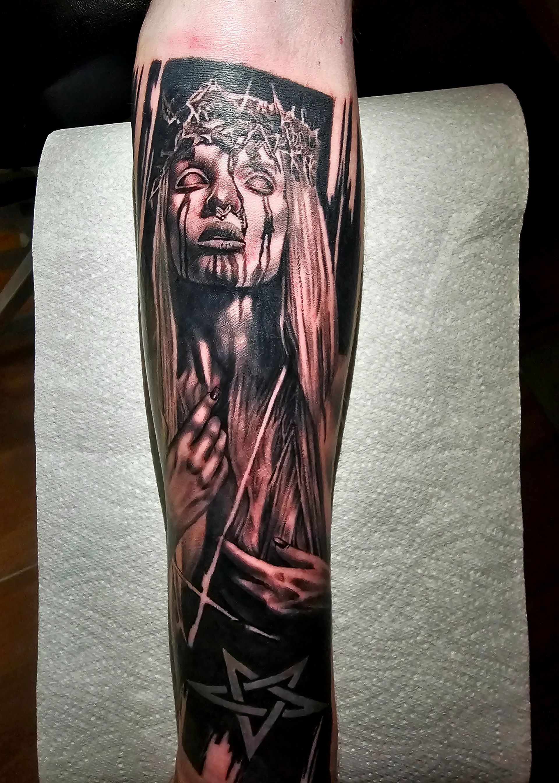 A black-and-grey tattoo on a forearm depicting a figure in a crown of thorns with weeping eyes and a pentagram below.