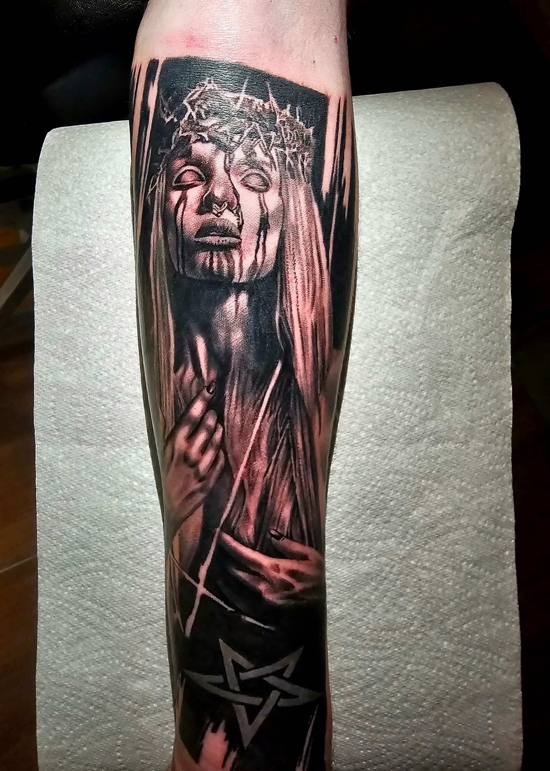 A dark, detailed forearm tattoo of a figure wearing a crown of thorns, with dripping lines and a pentagram at the base.