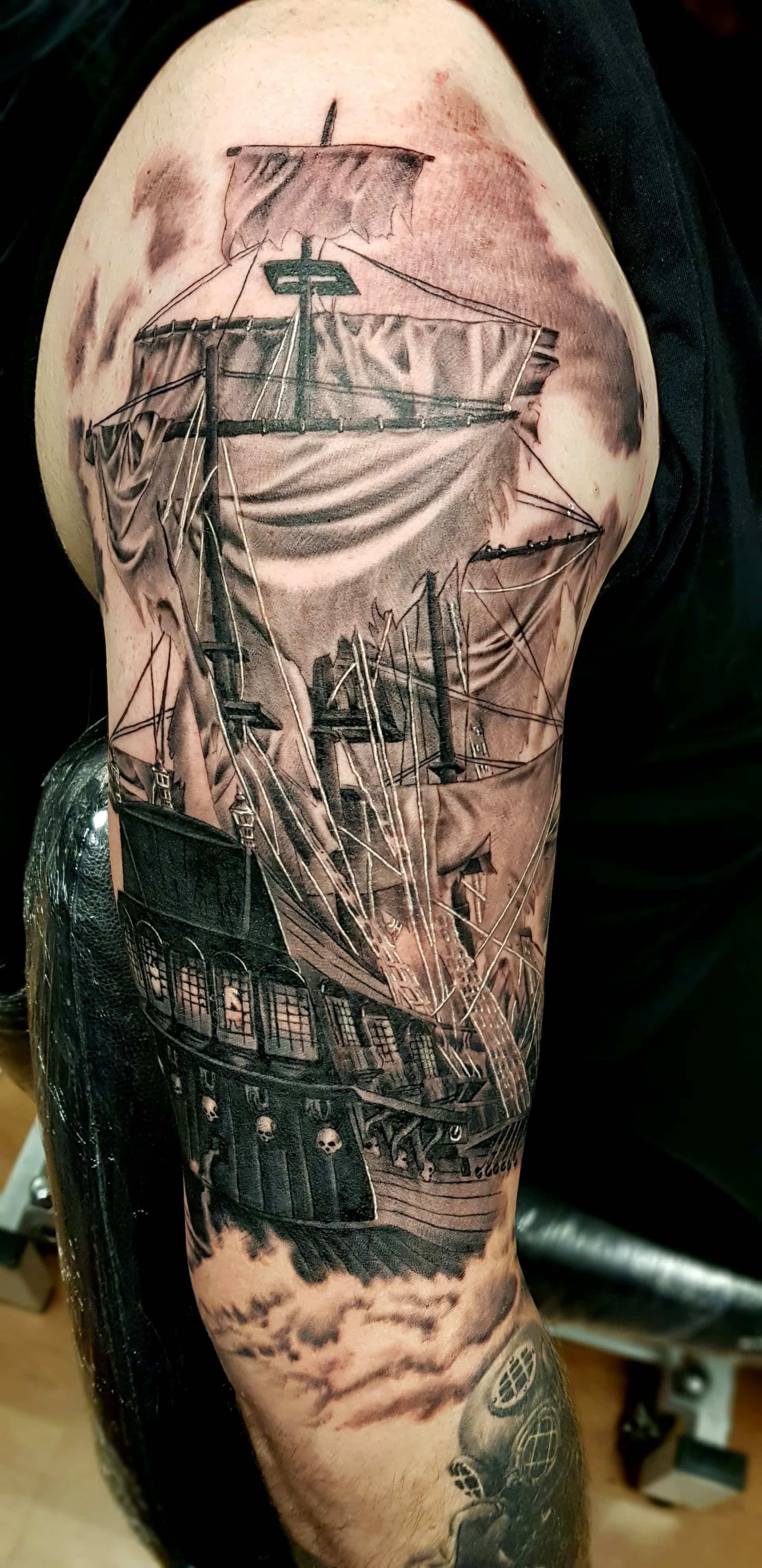 A black and grey tattoo on an upper arm featuring a detailed tall ship at sea, surrounded by stylized clouds and shading.