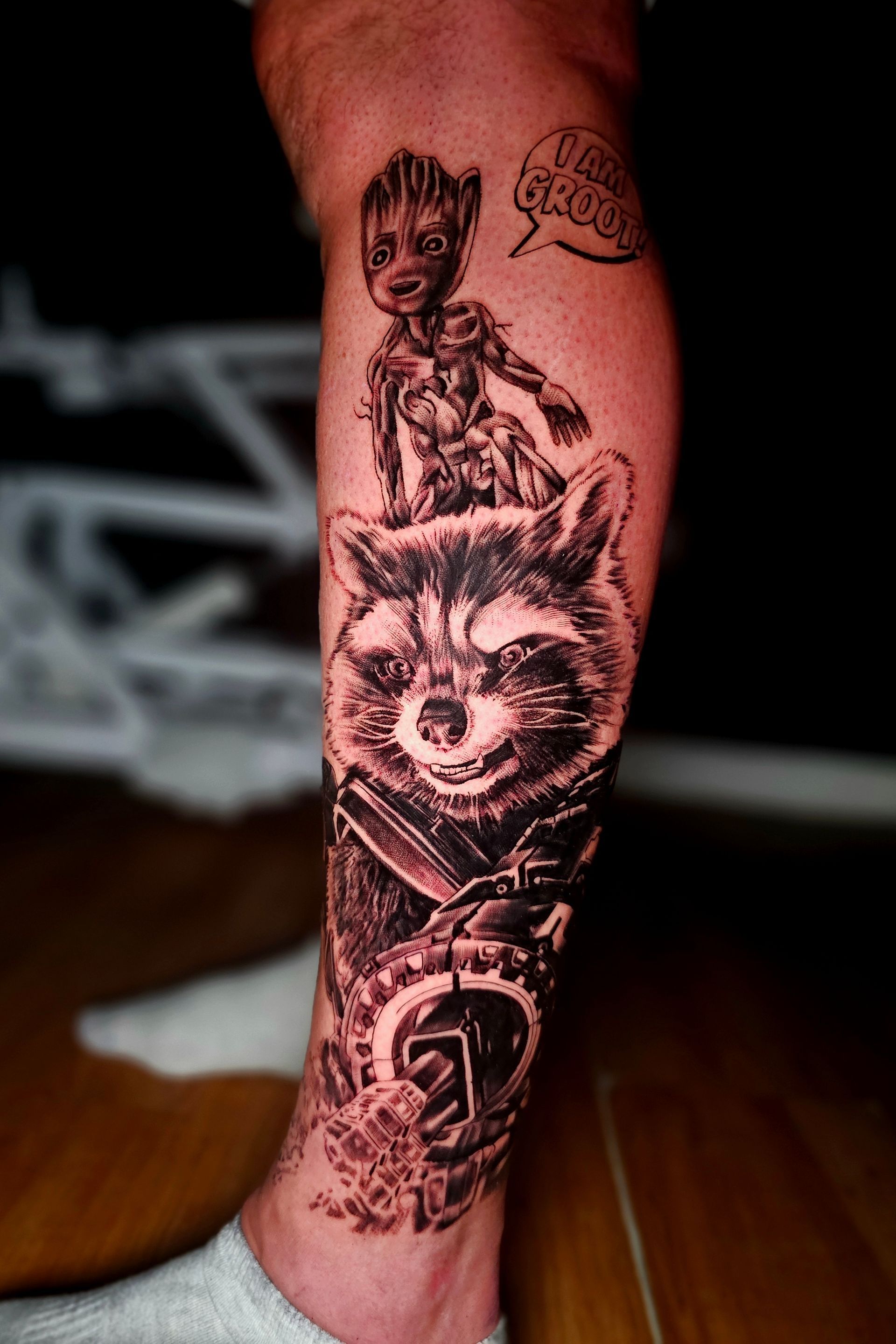 A black-and-grey tattoo on a lower leg featuring Rocket Raccoon and Baby Groot with a speech bubble saying 