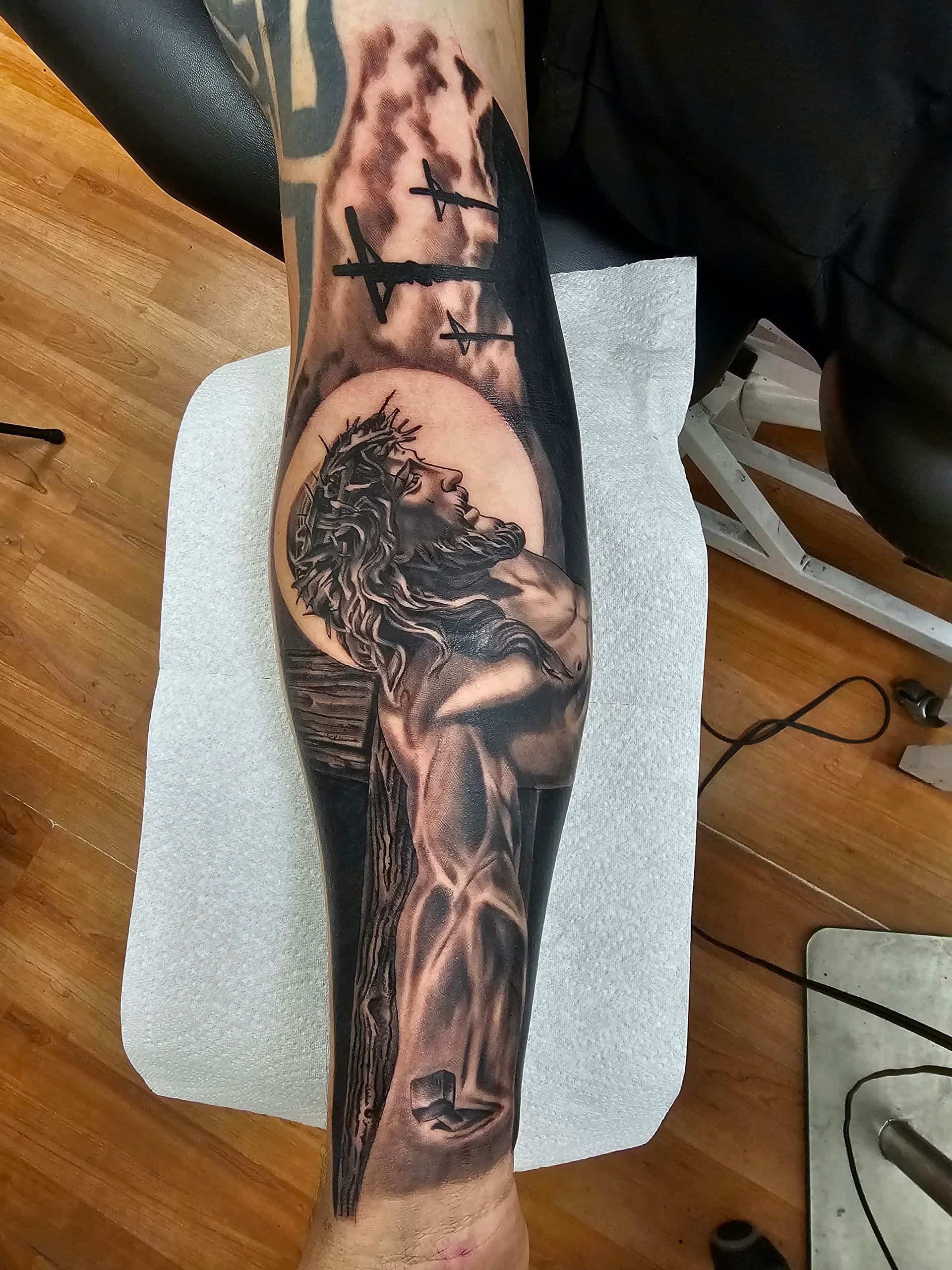A black and grey tattoo on a forearm depicting Jesus with a crown of thorns, set against three crosses on a hill.