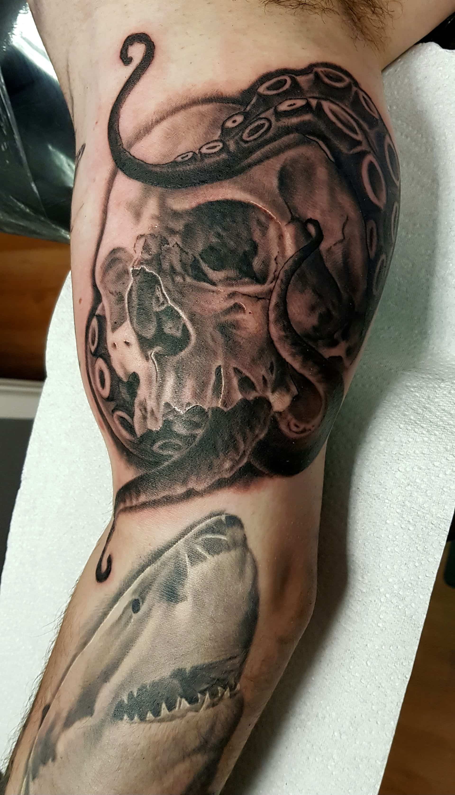 Black and gray tattoo on an arm showing an octopus tentacle wrapped around a human skull above a shark.