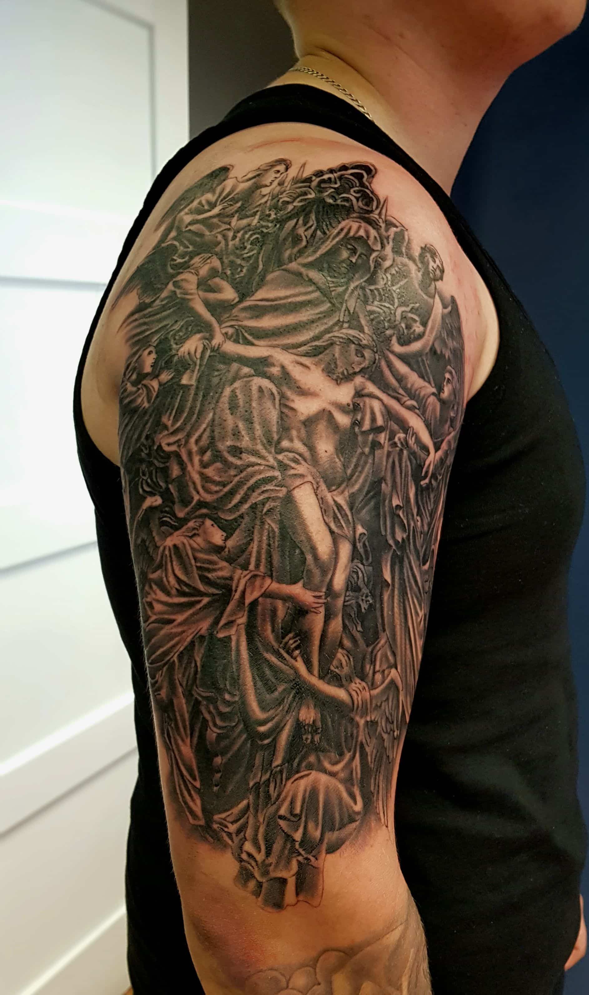 A close-up of a detailed, black-and-grey illustrative tattoo on a person's upper arm depicting angelic figures and drapery.