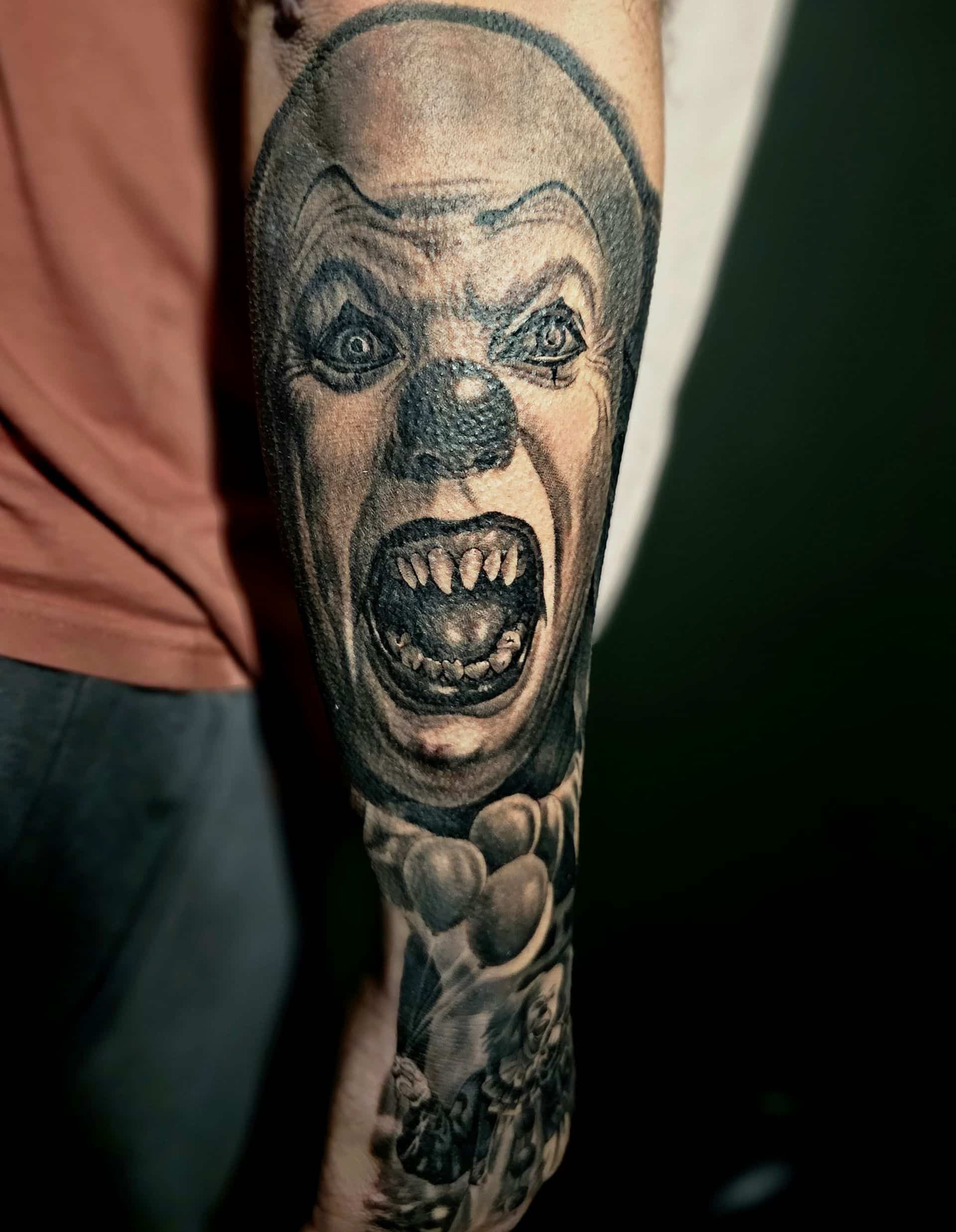 A forearm tattoo of a menacing, sharp-toothed clown face with a small cluster of balloons at the bottom.