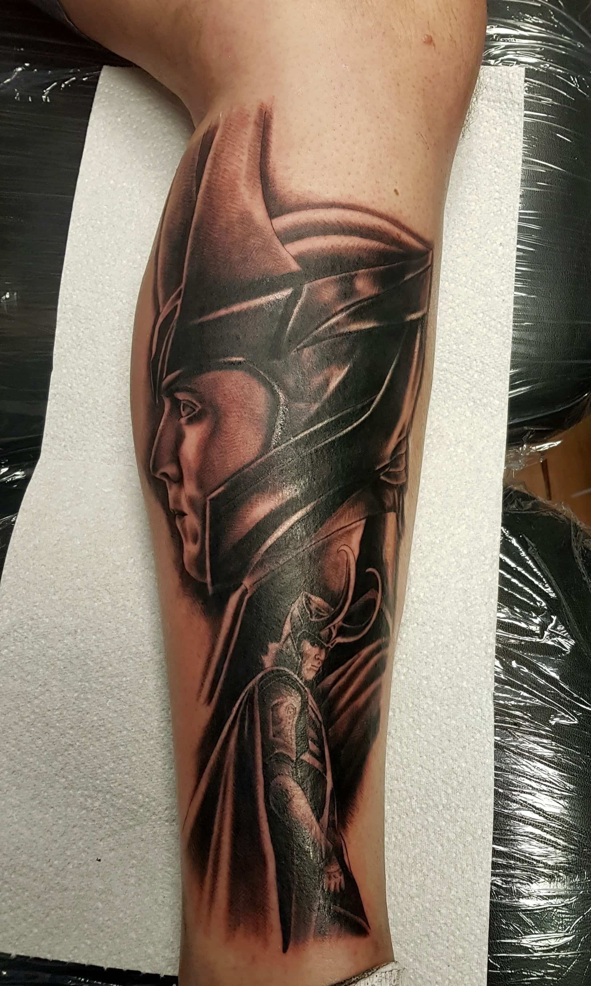 A black and grey tattoo on a person's calf featuring a detailed profile portrait of Loki in his horned helmet.