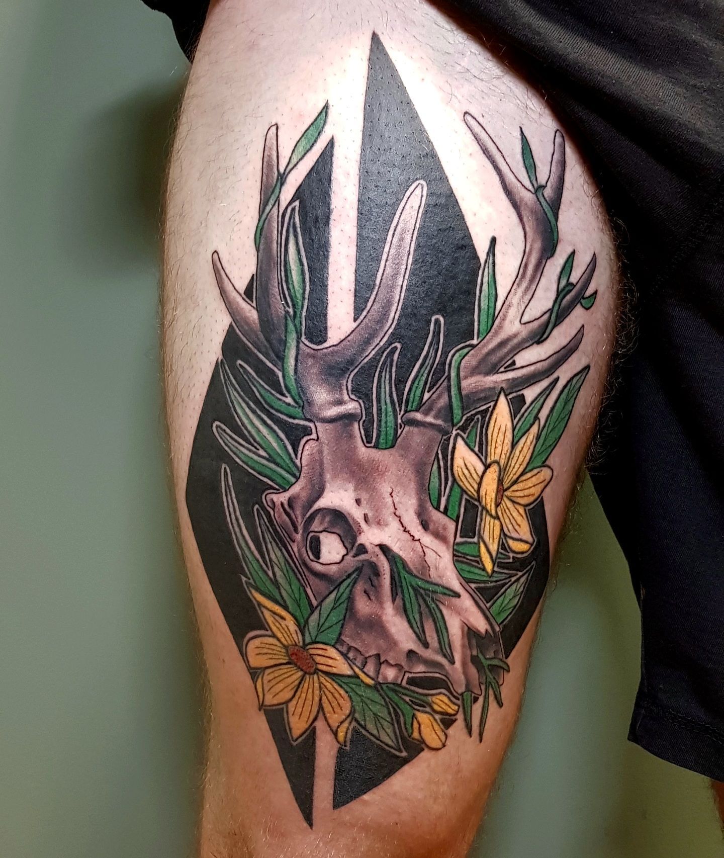 A tattoo on a leg featuring a deer skull intertwined with green foliage and yellow flowers against a black diamond shape.