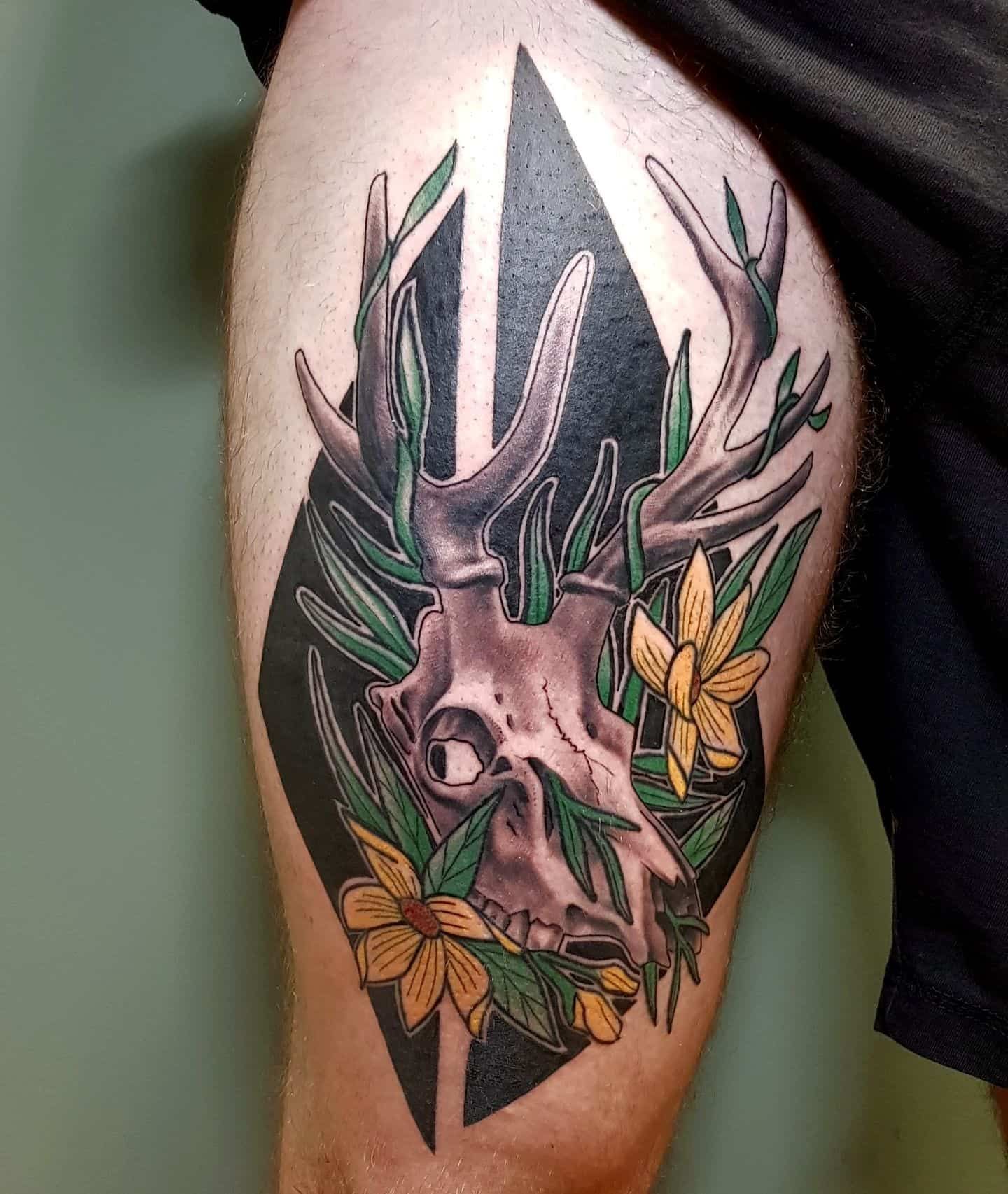 A tattoo on a leg featuring a deer skull surrounded by green leaves and yellow flowers against a black diamond background.