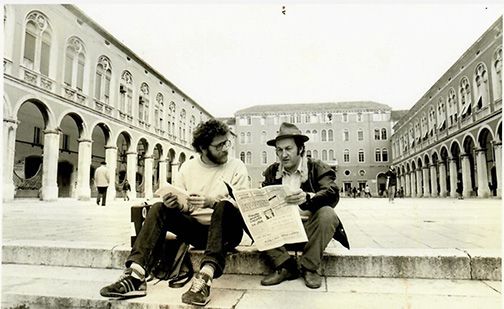 Two men are sitting on the steps of a building reading a newspaper.